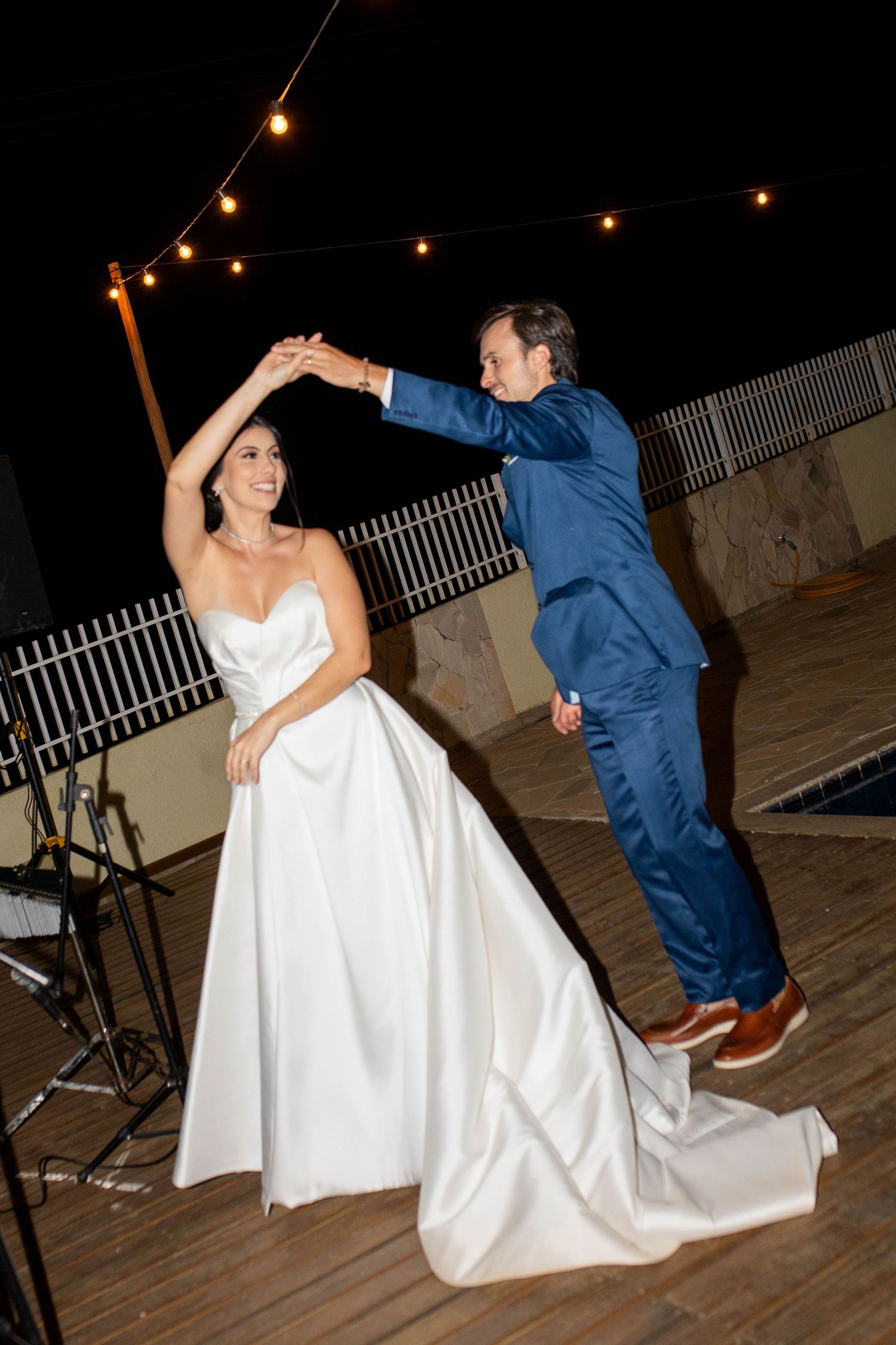 A groom in a blue suit twirls his bride in a white gown during their first dance under string lights at night.