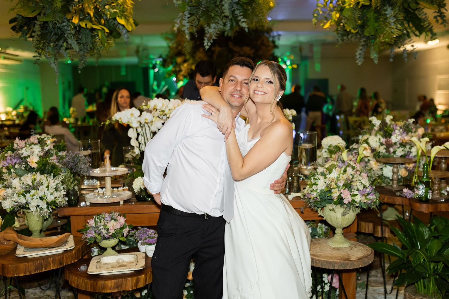 Bride and groom embracing and smiling at their elegant wedding reception with floral arrangements and green lighting.