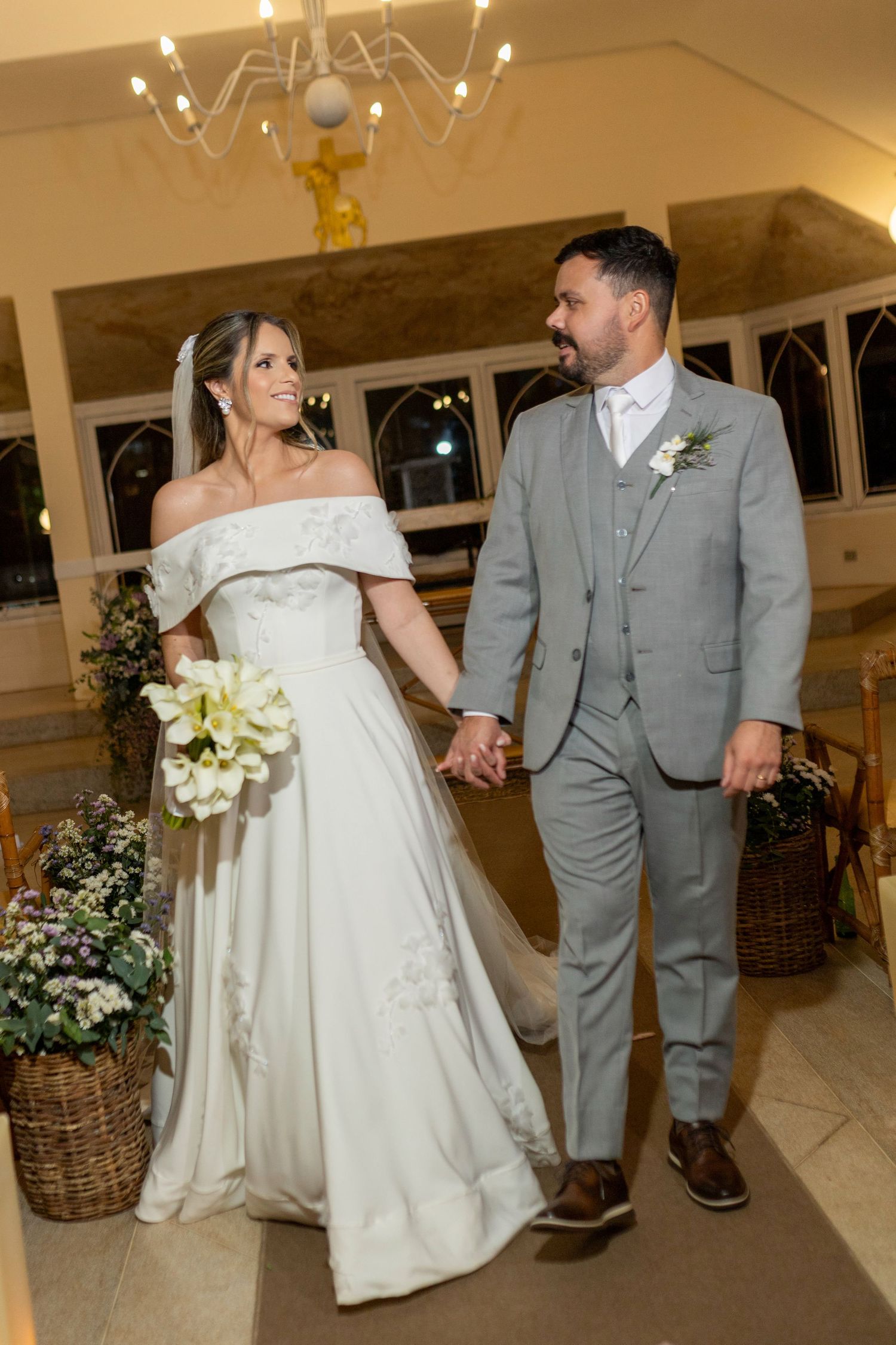 A bride in a white off-shoulder gown holds a bouquet while walking with a groom in a gray suit at their wedding ceremony.