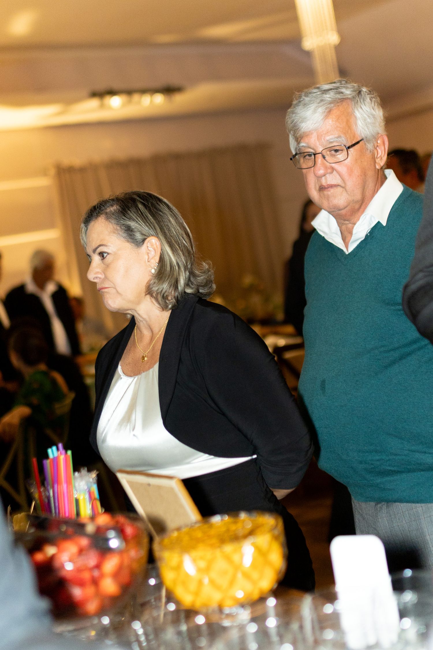 Older couple at a formal event standing near a buffet table with fruit bowls and colorful straws.