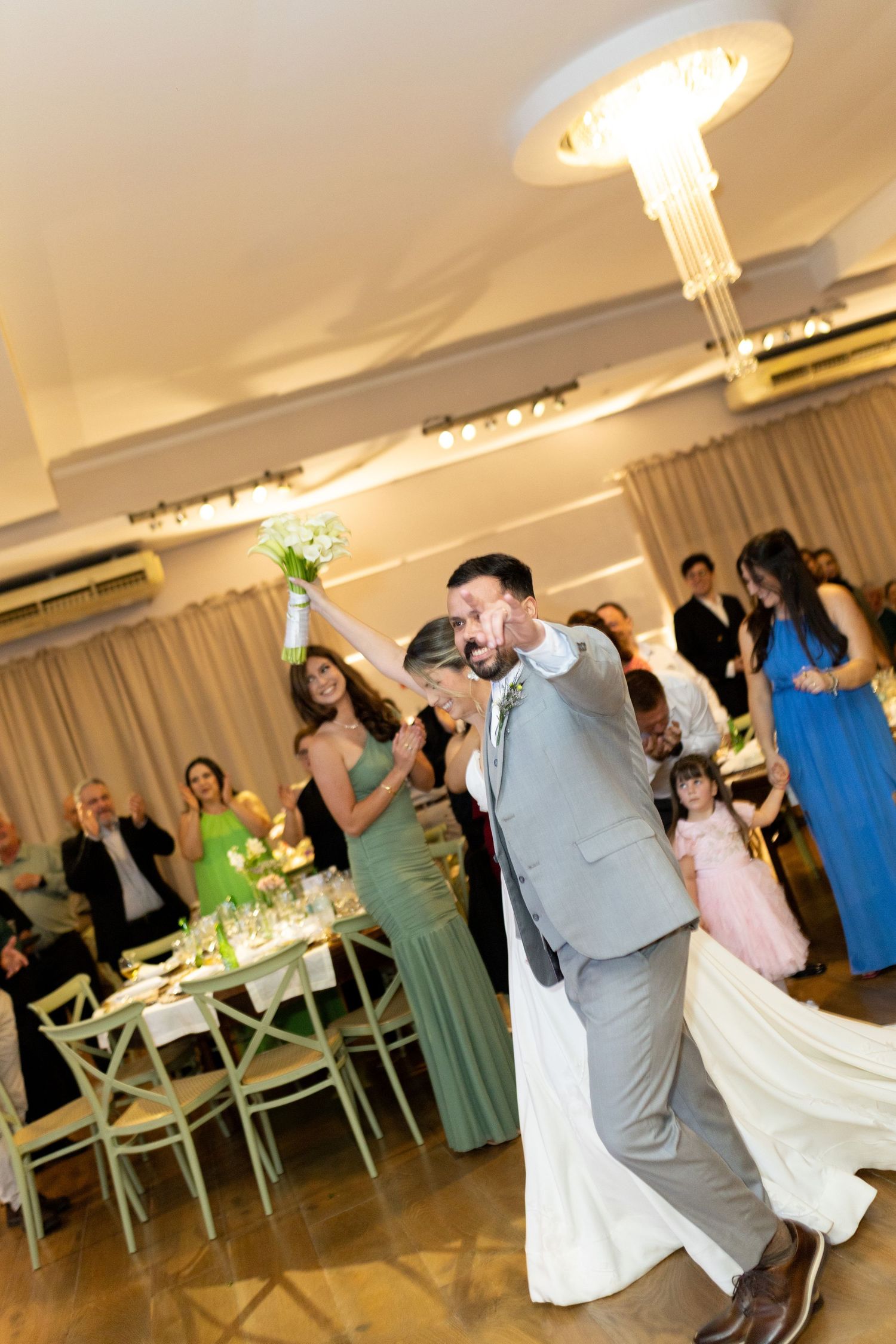 Bride and groom share first dance at elegant wedding reception, bride holding bouquet aloft, guests celebrating in background.