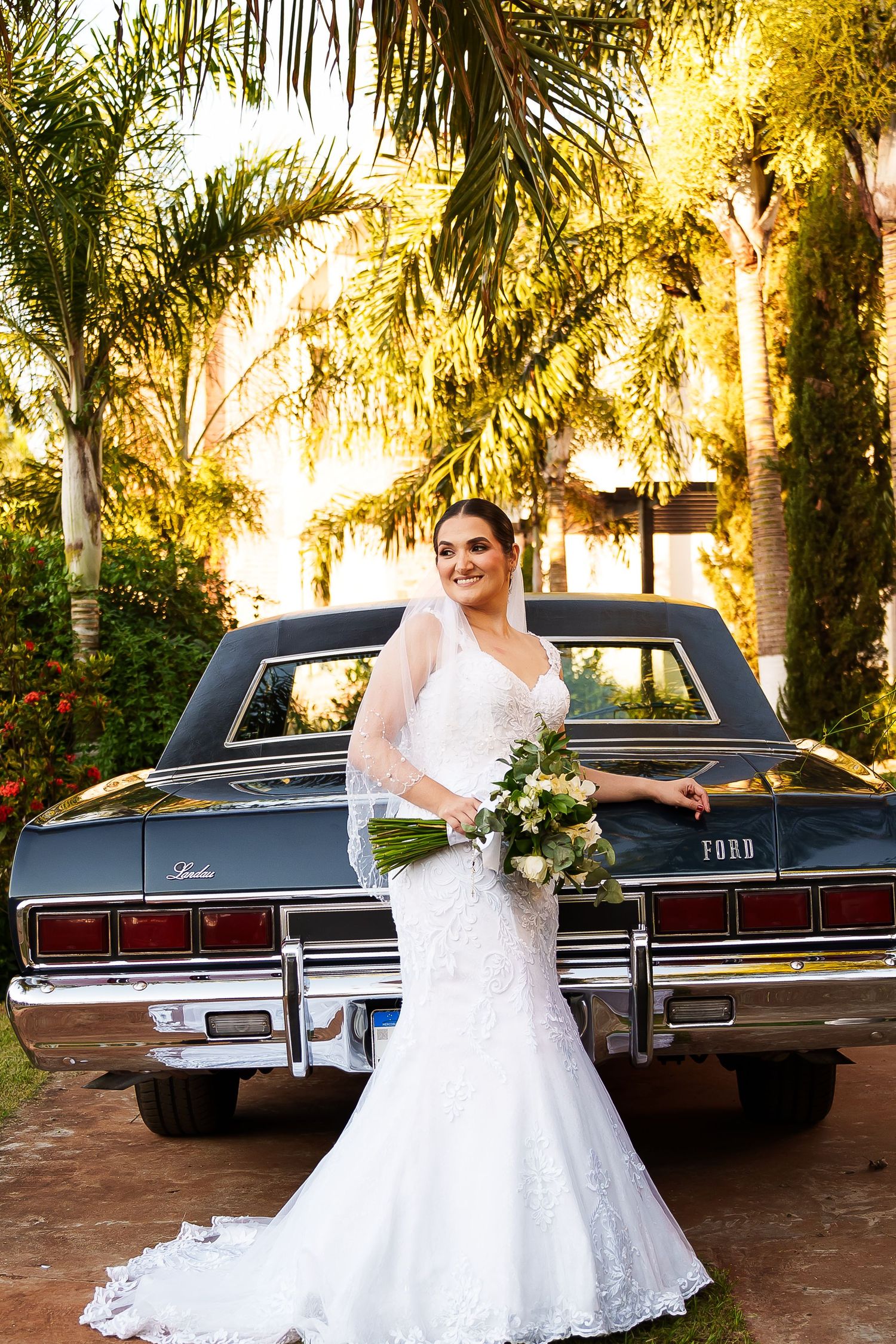 Bride in lace mermaid gown with veil holds bouquet beside vintage Ford Lincoln under tropical palm trees.