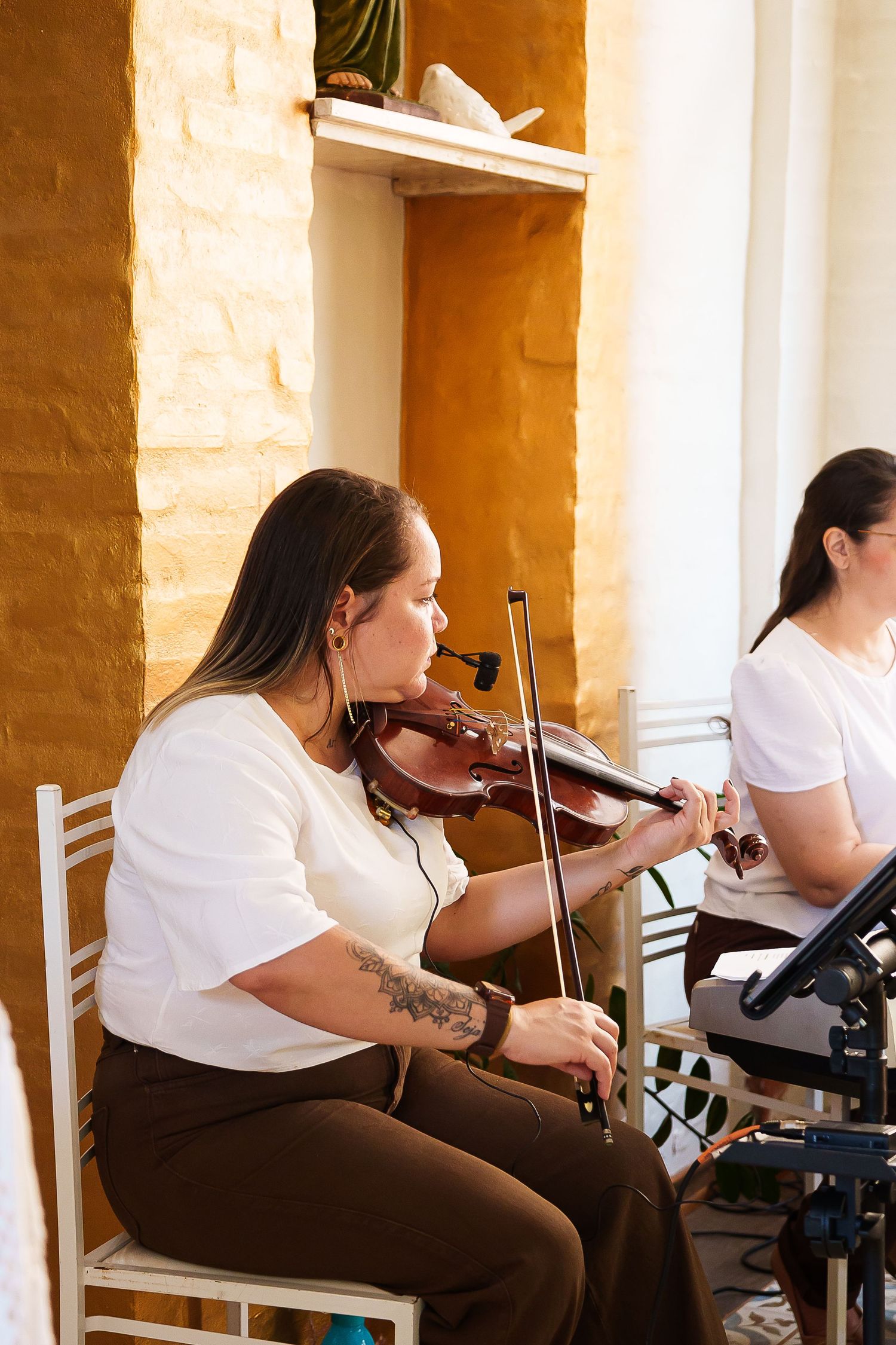 Woman in white top playing violin while seated, with another musician visible in the background indoors.