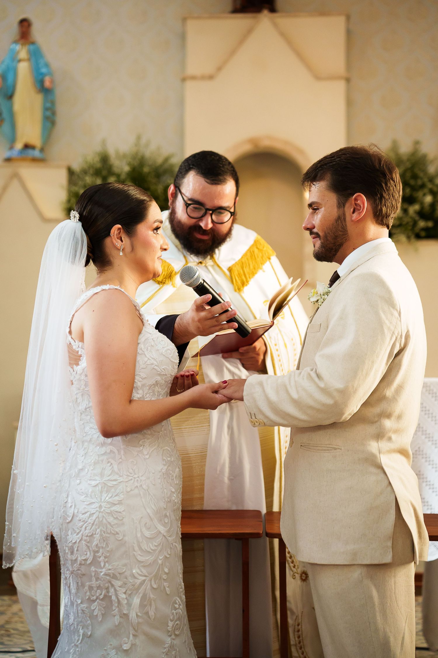 Bride and groom exchange vows at church altar with officiant holding microphone and book during wedding ceremony.