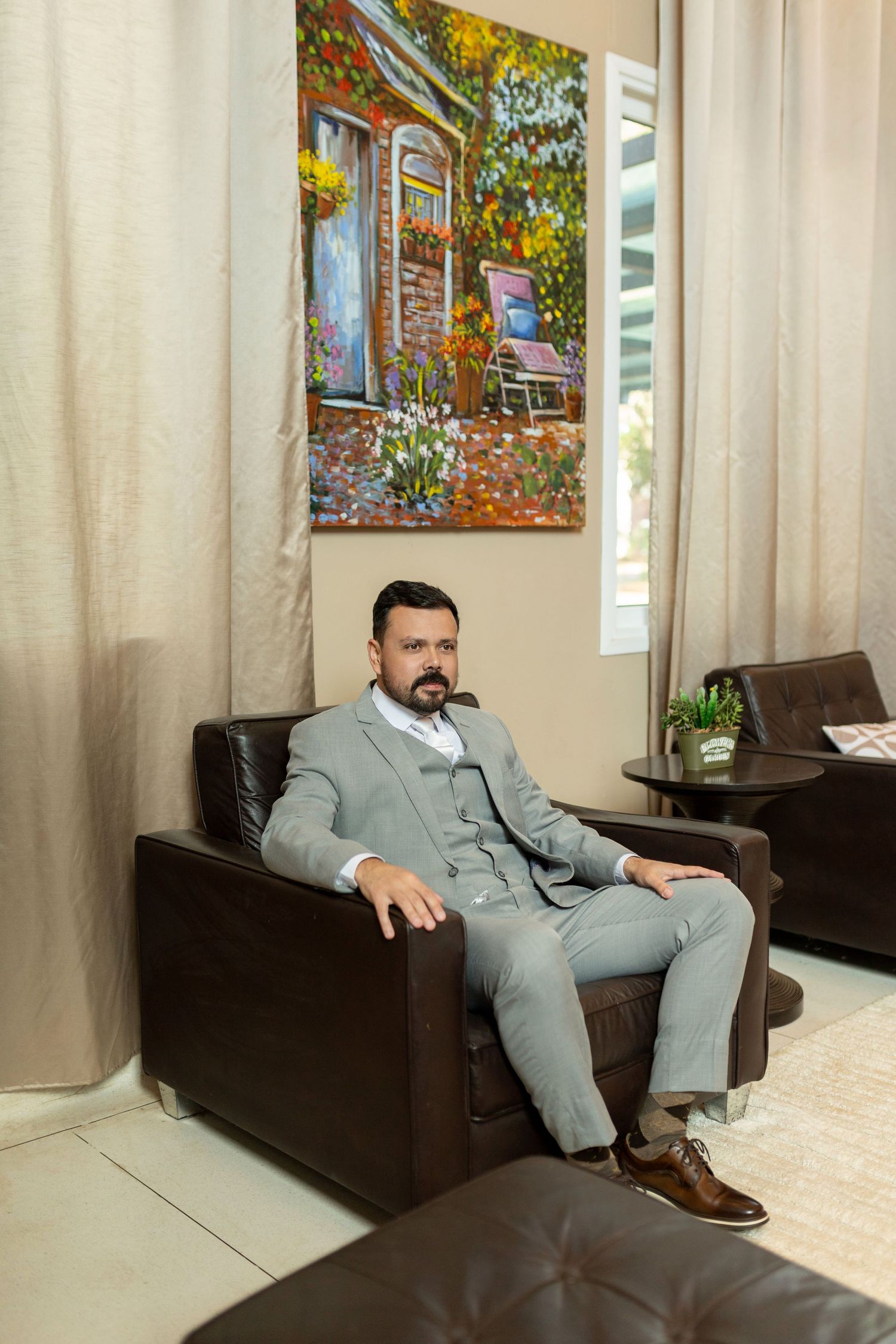 Bearded man in gray three-piece suit sitting in leather armchair beneath colorful painting.