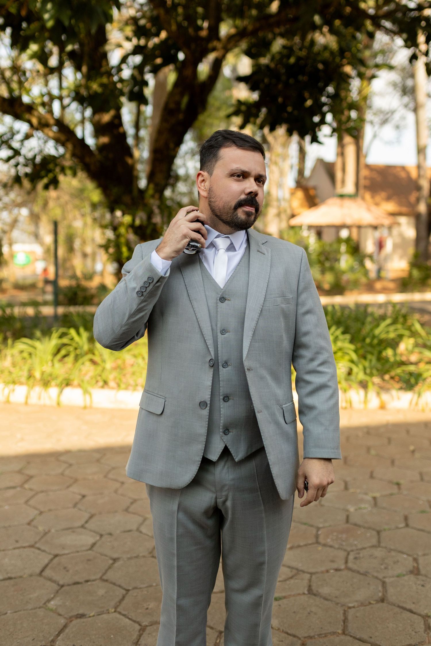 Man in gray three-piece suit standing outdoors in a park, holding a phone near his ear.