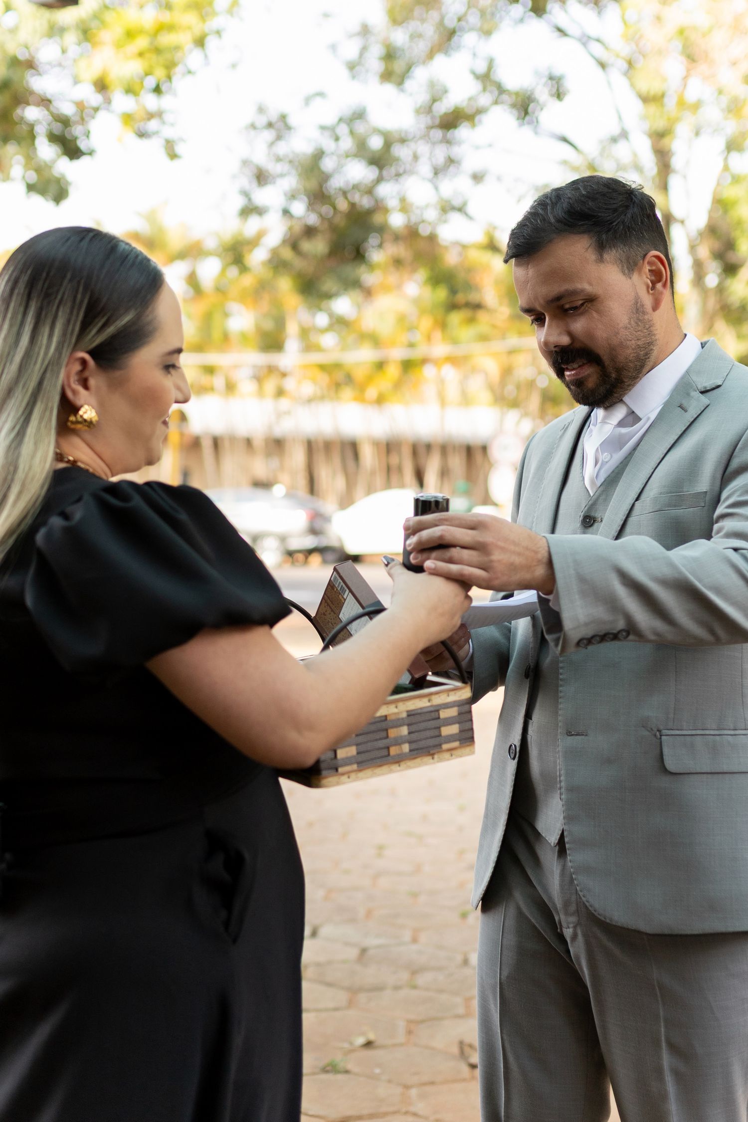 A woman in black dress and man in gray suit exchange rings outdoors at a wedding ceremony.