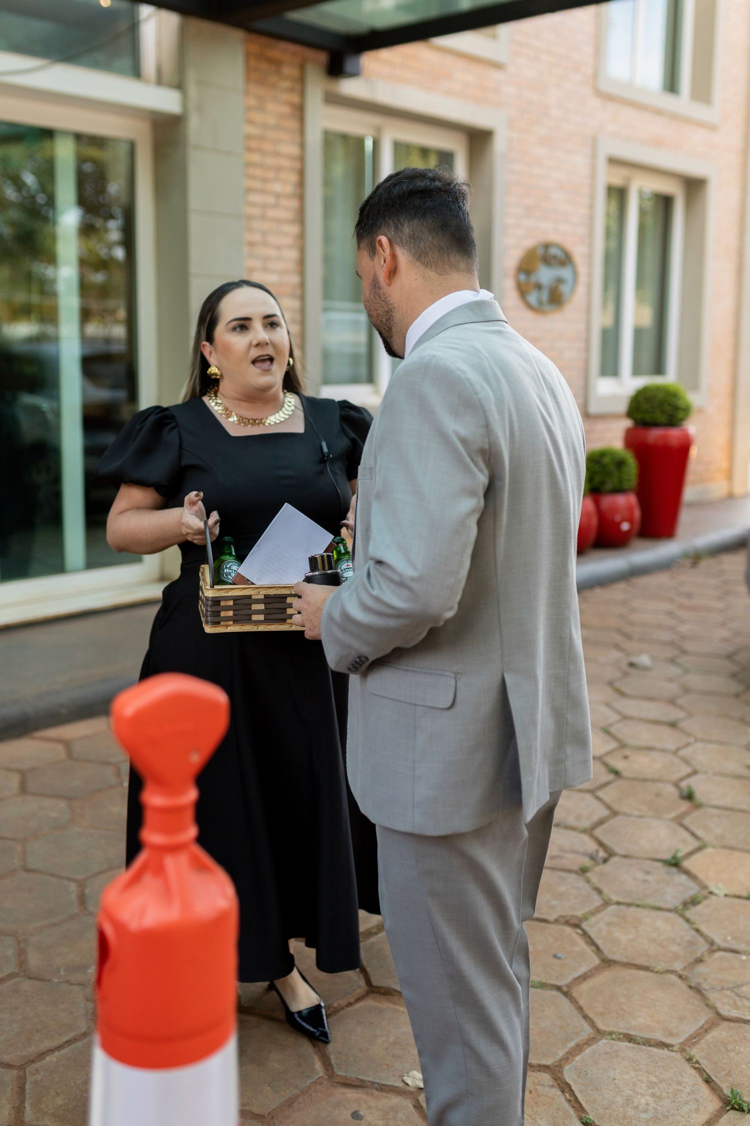 Woman in black dress holding basket chats with man in gray suit outside a brick building.
