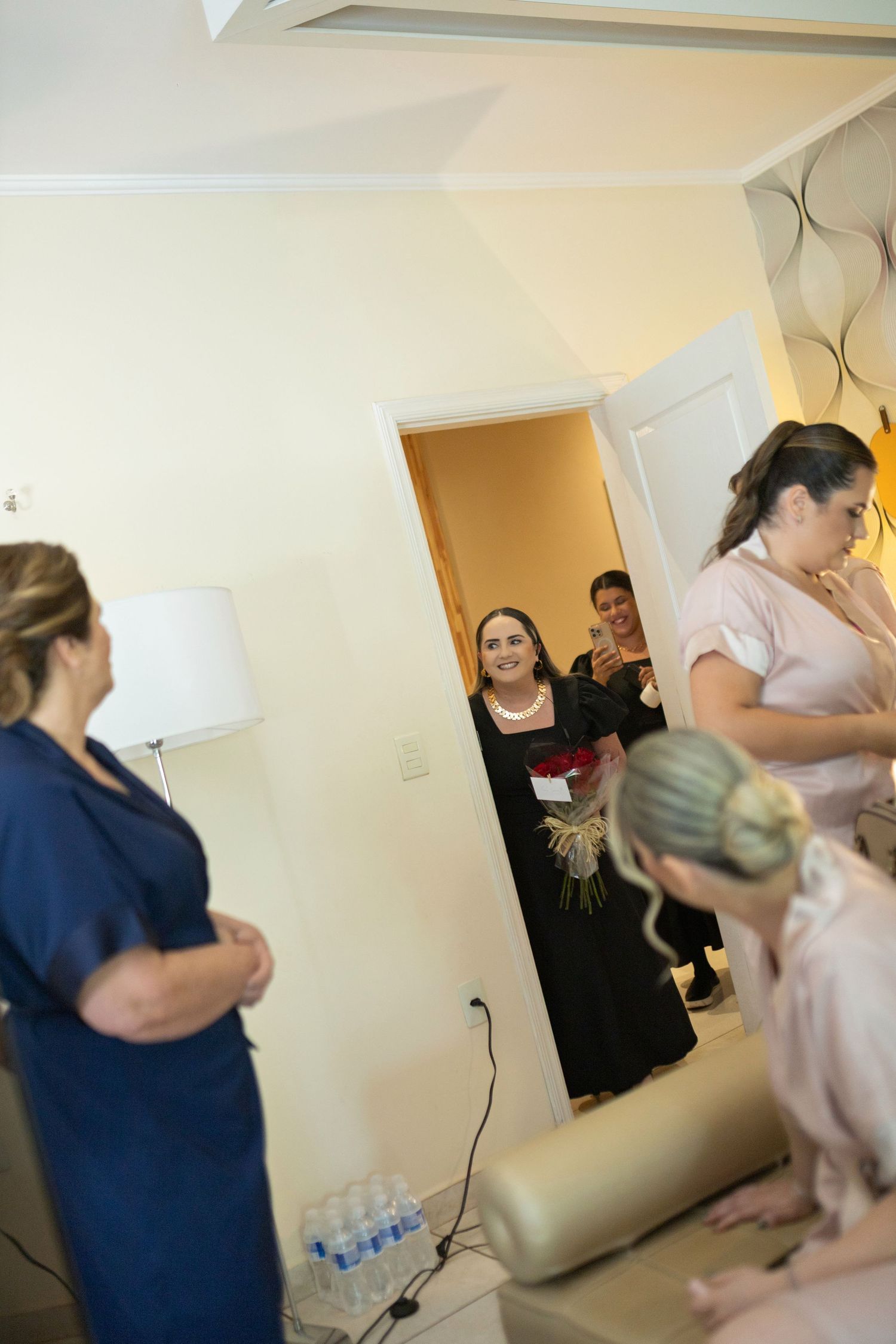 Bridesmaids in pink and navy gather in a hotel room as a smiling guest arrives holding a bouquet of flowers.
