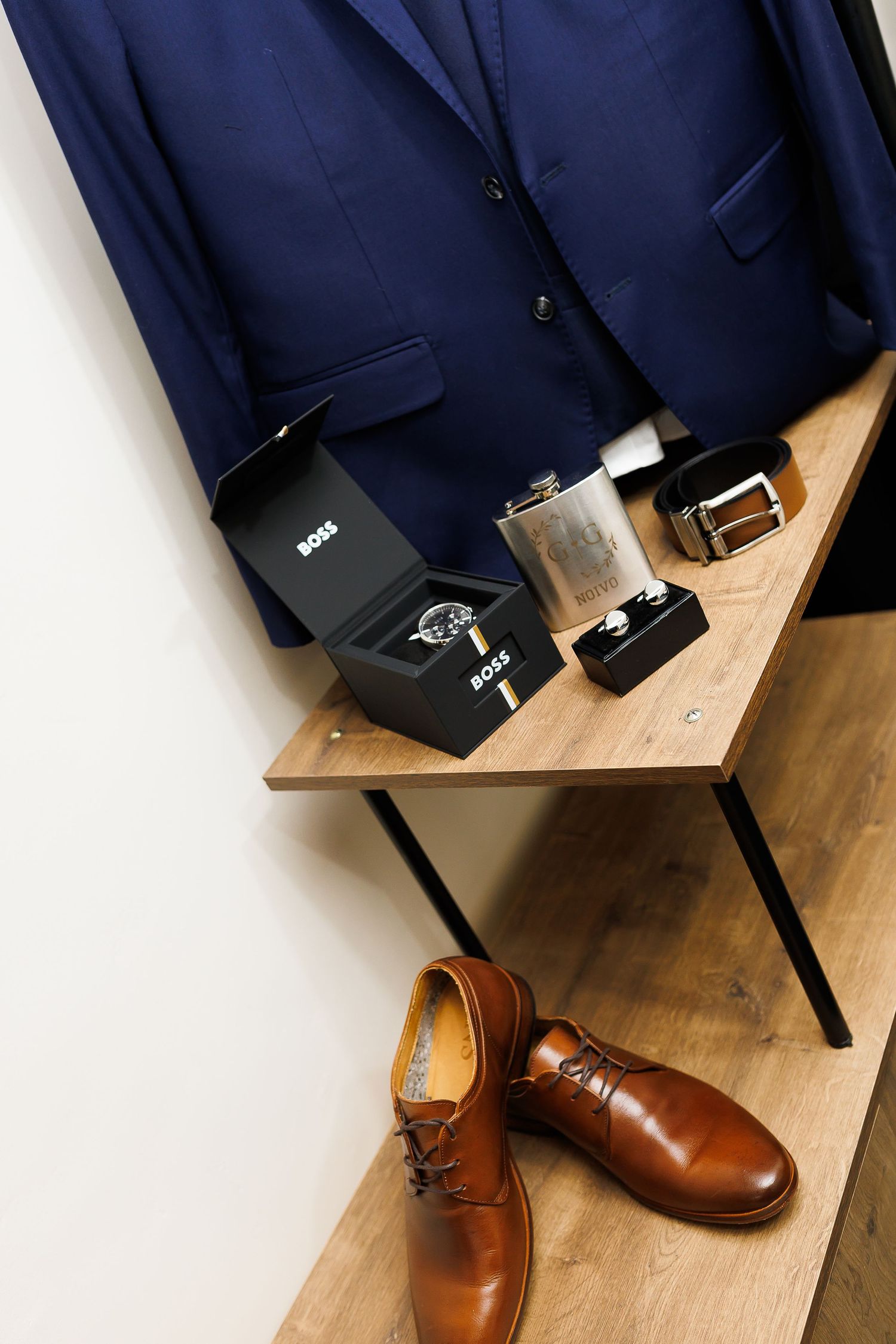 Navy blue suit with Hugo Boss watch box, flask, belt, cufflinks on wooden shelf, and tan leather Oxford shoes below.