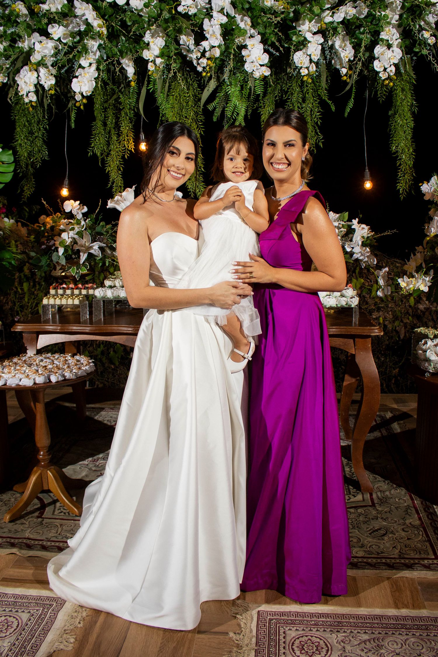 Bride in white gown and woman in purple dress holding a toddler at an elegant floral wedding reception.