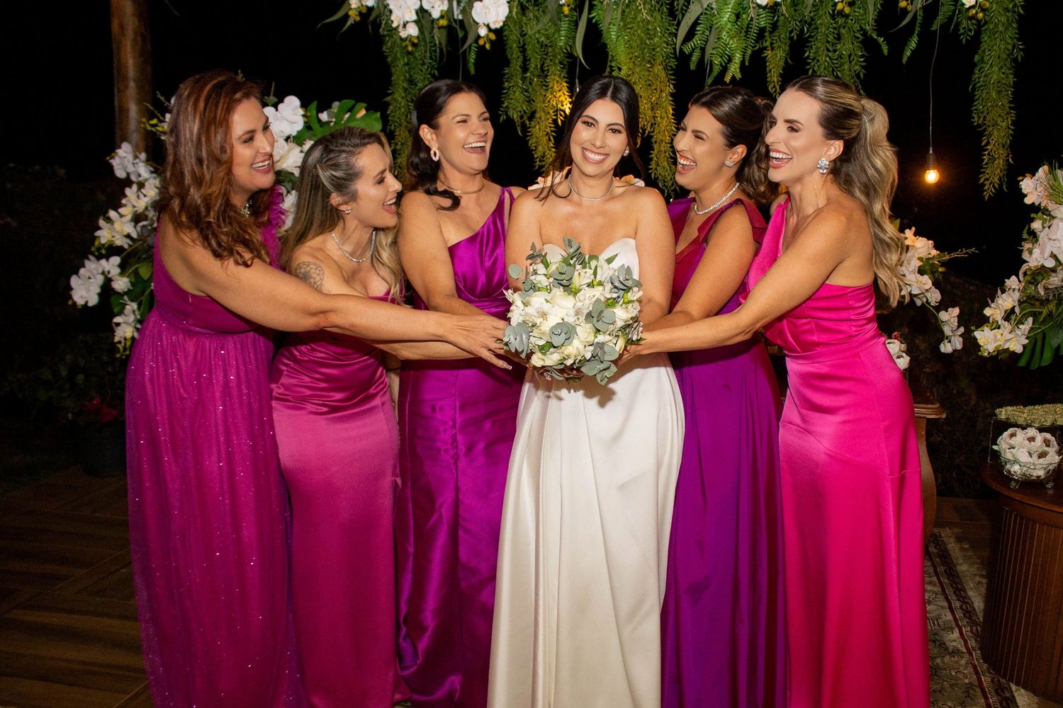 Bride in white gown holds bouquet surrounded by bridesmaids in pink and magenta satin dresses at outdoor wedding.