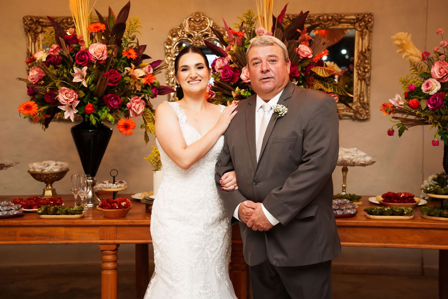 Bride in white lace gown poses with older man in gray suit before floral arrangements at wedding reception.