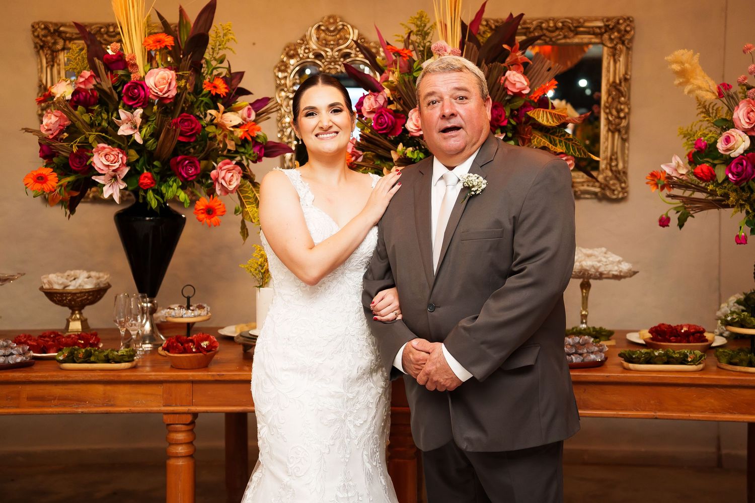 Bride in white lace gown poses with older man in gray suit before floral arrangement at wedding reception.