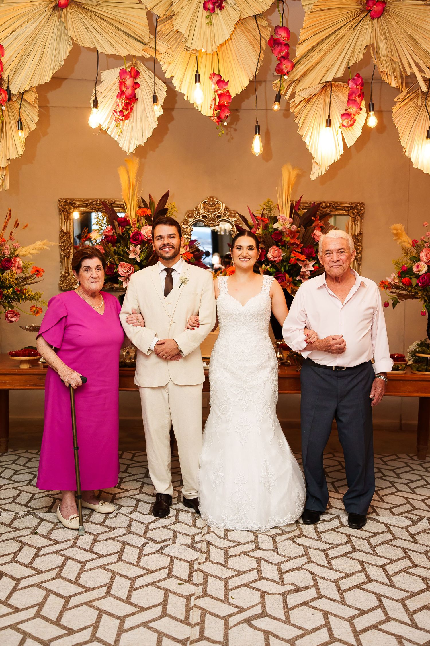Bride and groom pose with elderly parents at floral wedding reception with tropical palm leaf decorations.