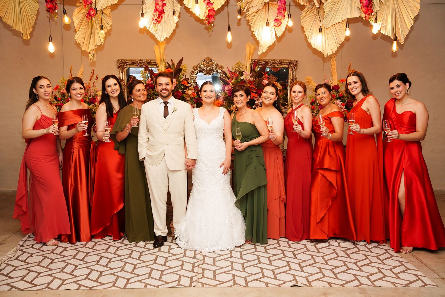 Wedding party posing together, bride and groom centered, bridesmaids in red and green gowns, elegant floral decor backdrop.