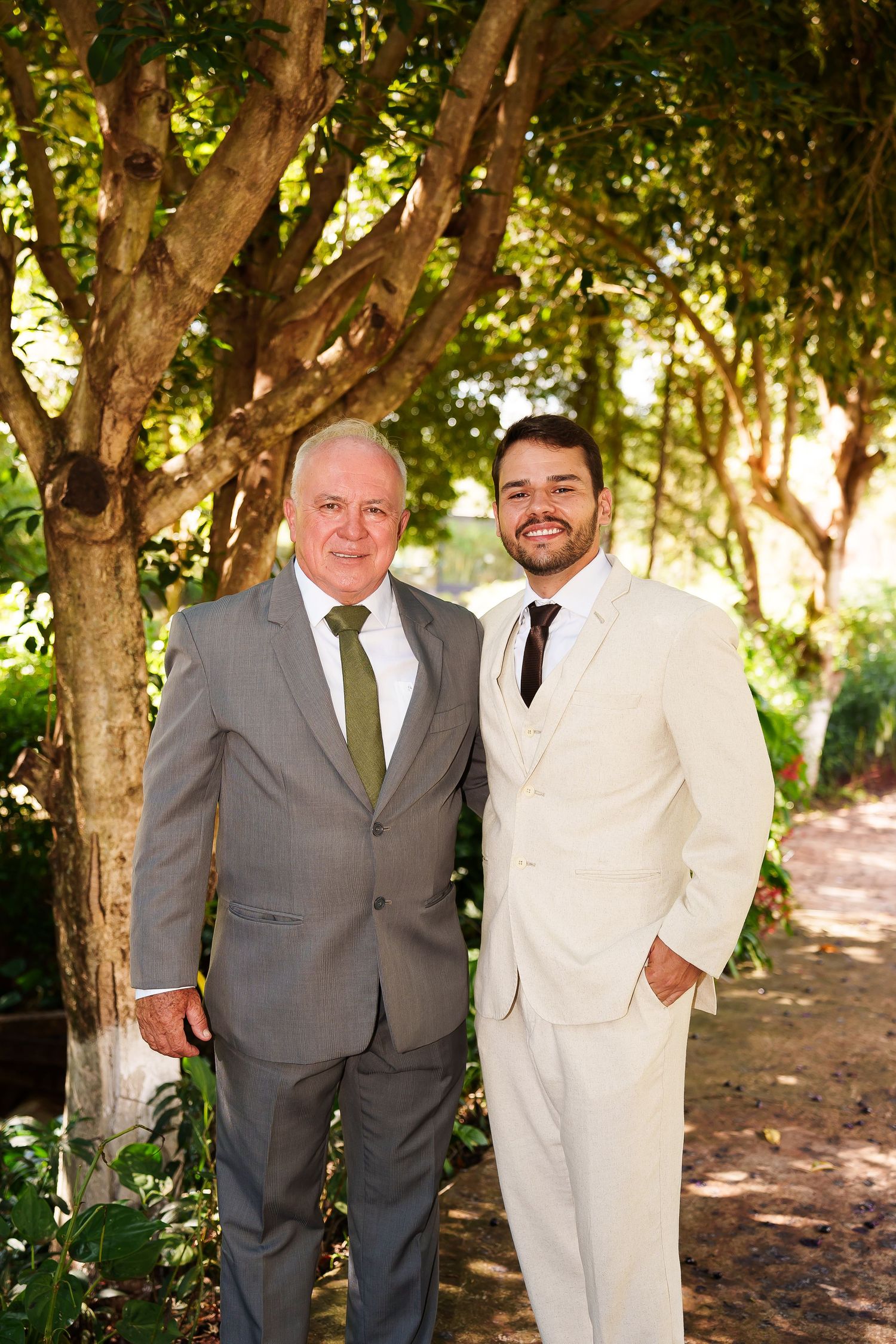 Two men in suits posing outdoors at a wedding, surrounded by lush green trees on a sunny day.