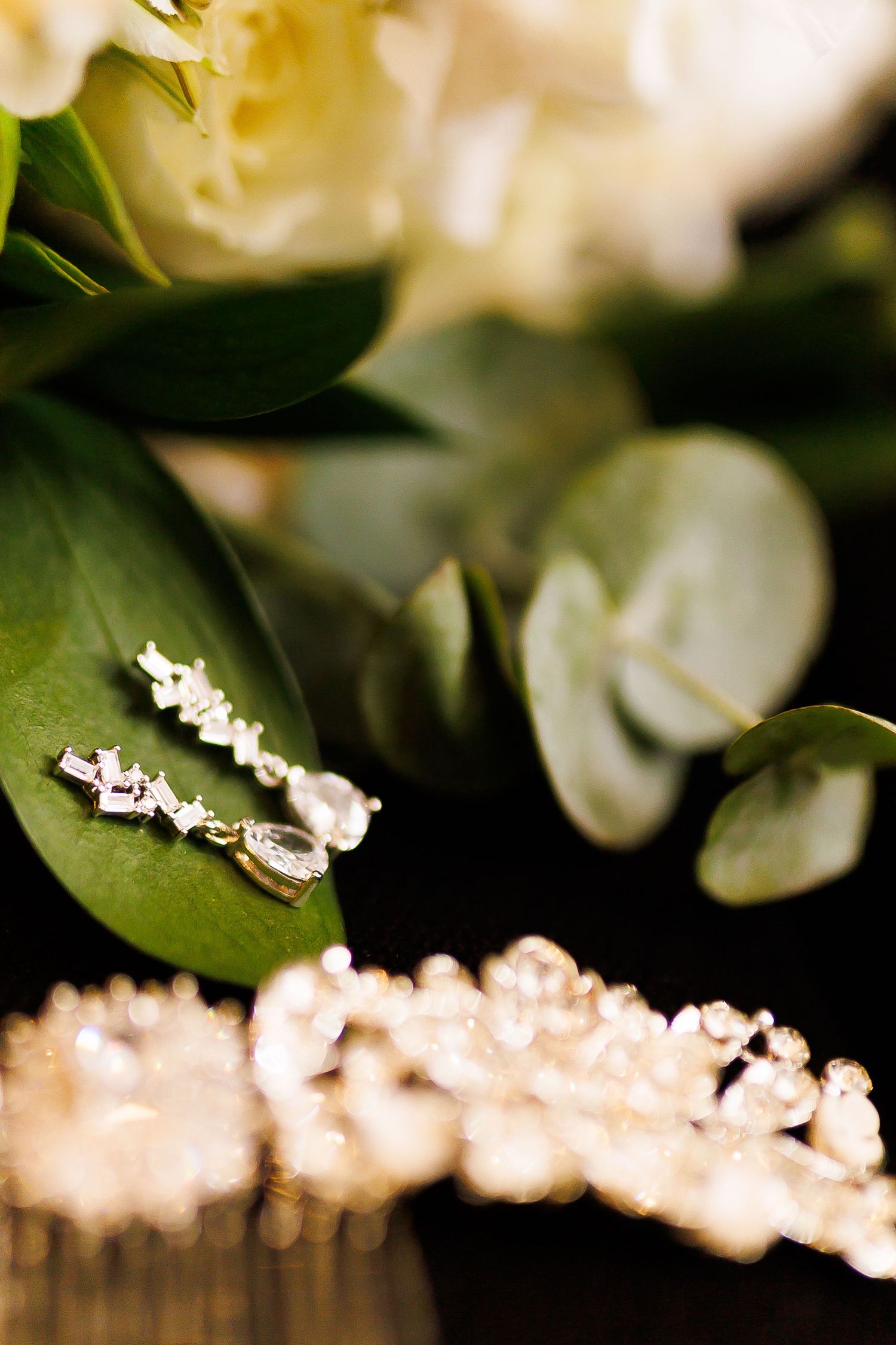 Elegant crystal drop earrings resting on green eucalyptus leaves beside white flowers and a sparkling rhinestone bridal headpiece.