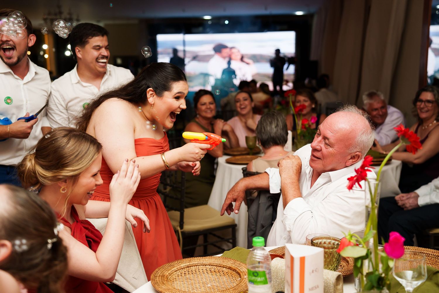 Joyful wedding reception guests laughing and celebrating, woman in red dress playfully interacting with older man at festive party.