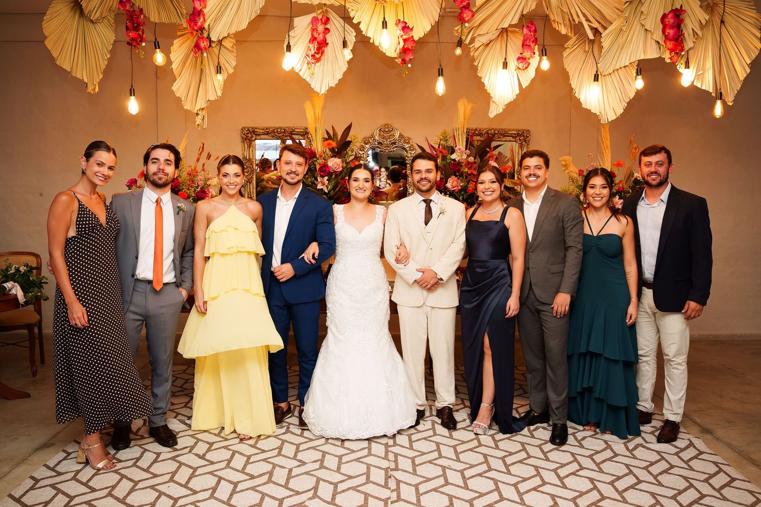 Wedding party posing together in elegant venue with floral ceiling decor, bride in white gown surrounded by guests in formal attire.