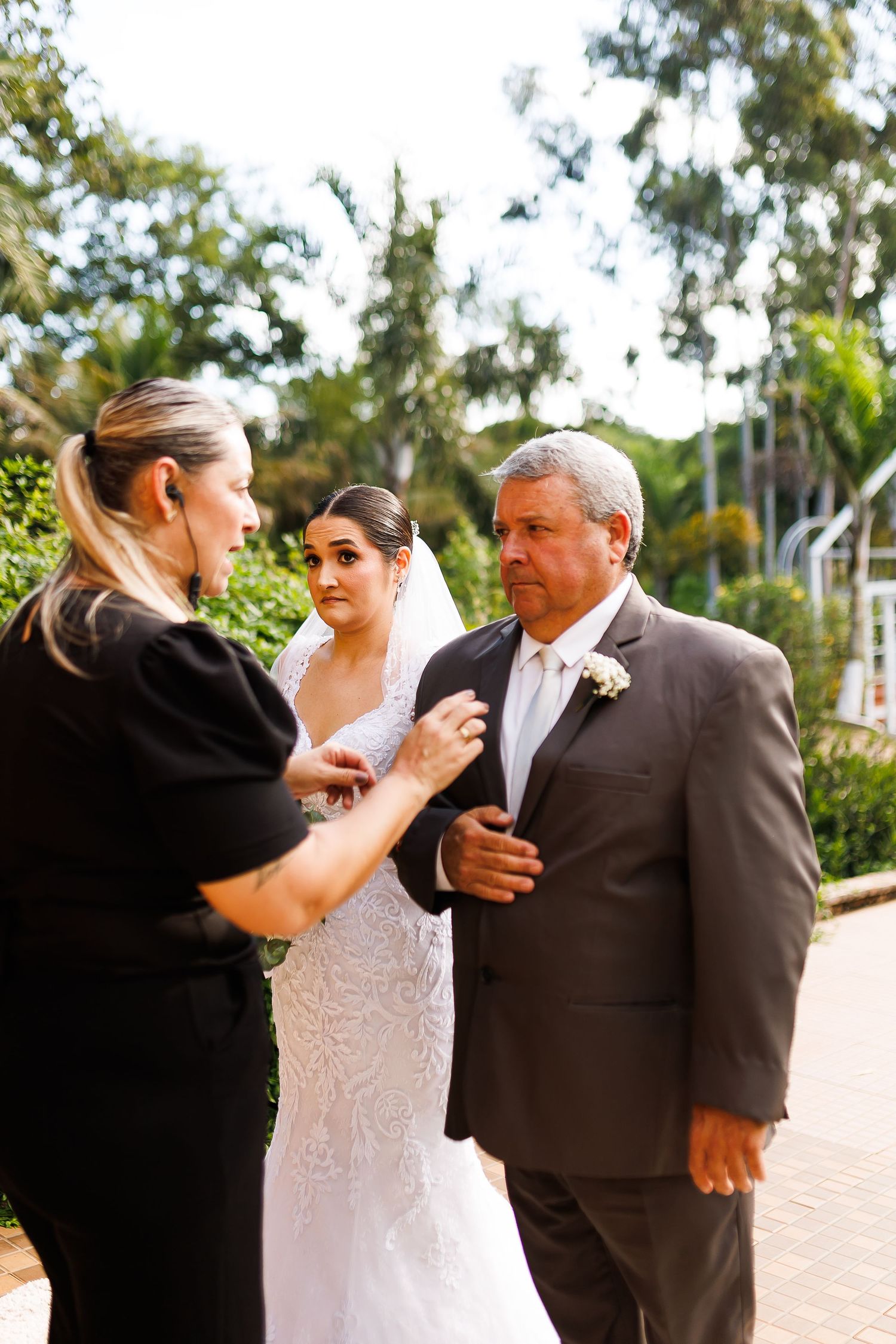 Bride in white lace gown stands with groom in gray suit as wedding coordinator adjusts boutonniere outdoors.