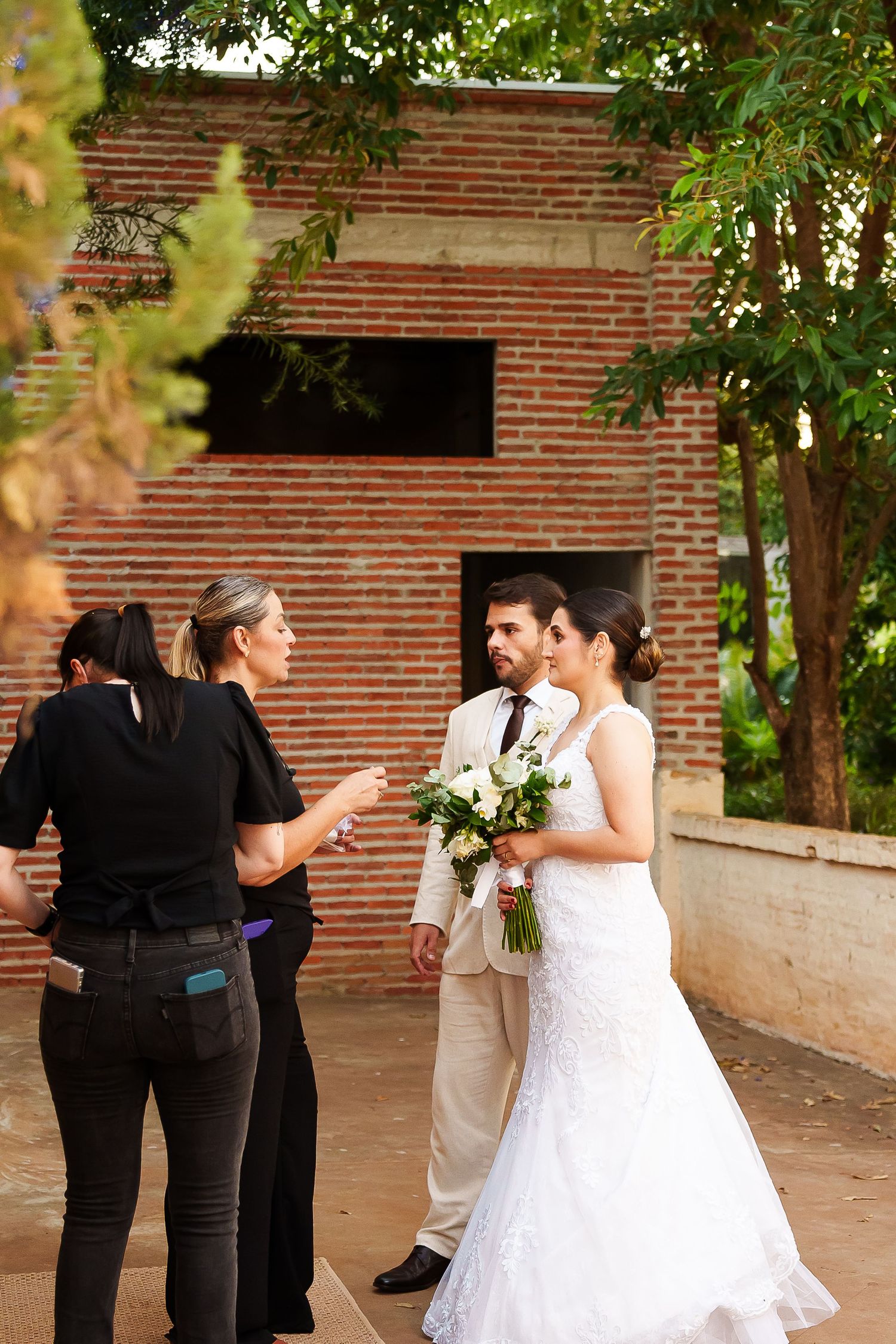 Bride in white gown holding bouquet stands with groom in tan suit, speaking with wedding coordinator outdoors.