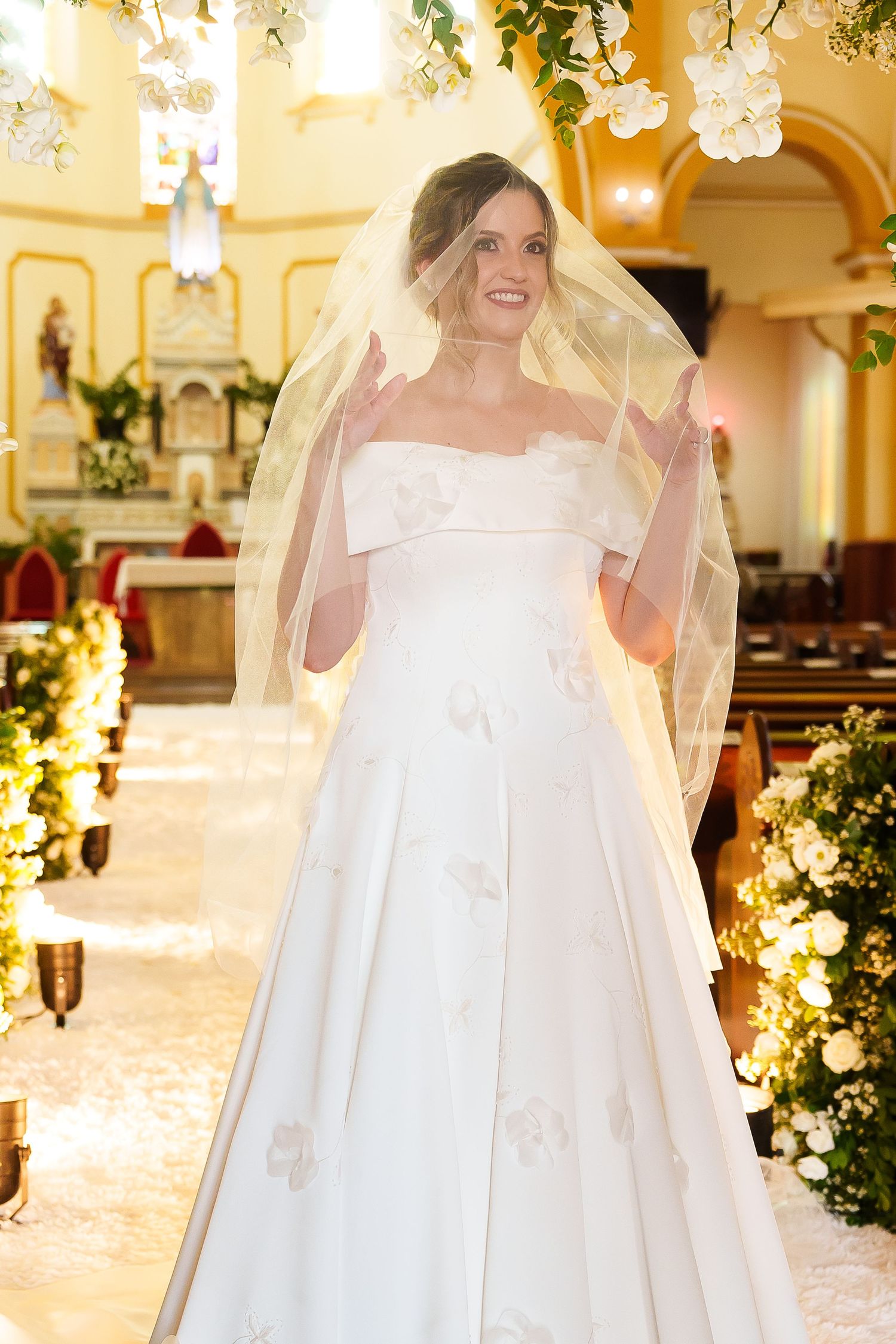 Smiling bride in white off-shoulder gown with veil lifted, standing in decorated church aisle with floral arrangements.