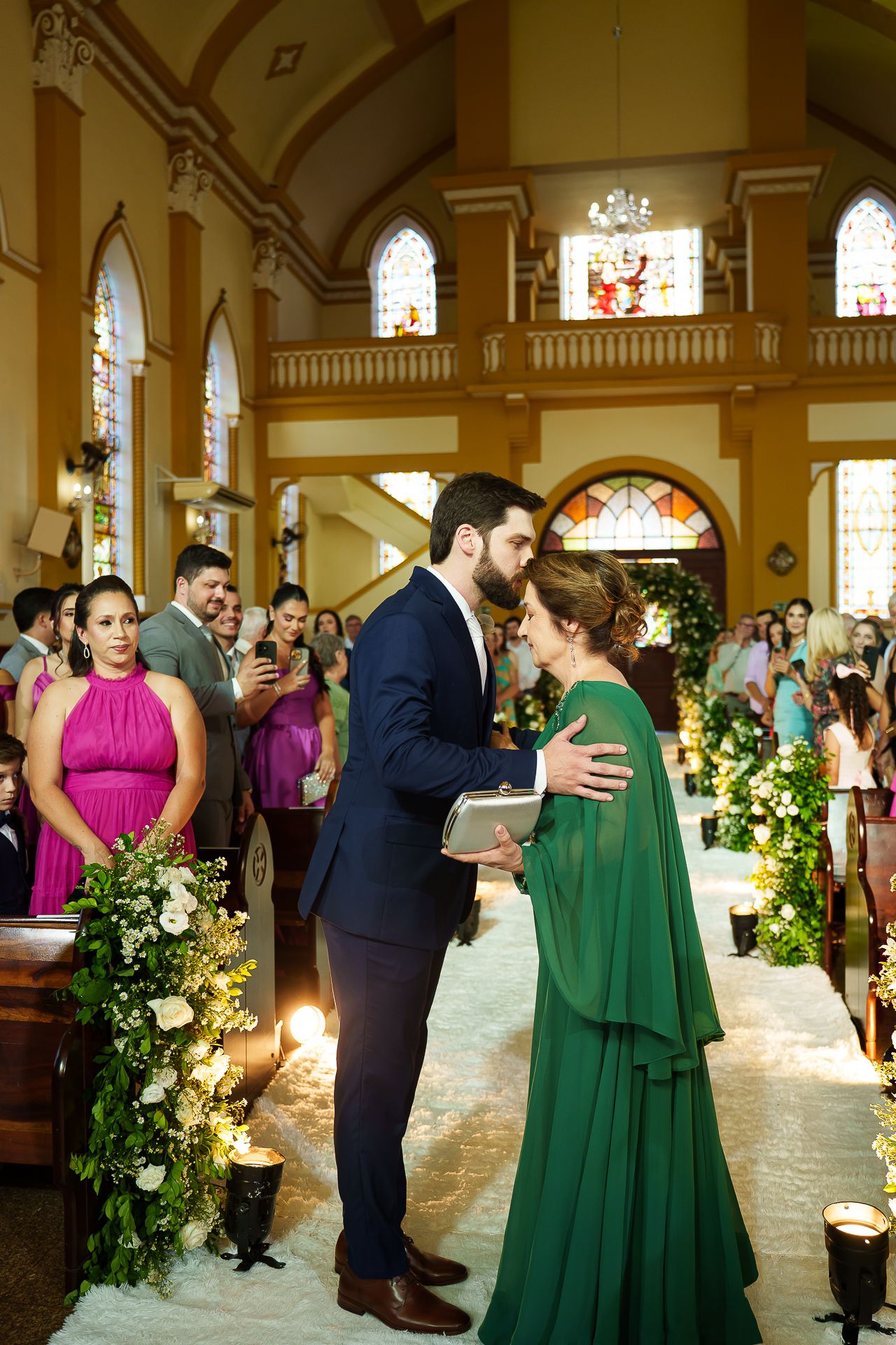 Groom in navy suit greets woman in green cape dress at church wedding ceremony aisle with floral decorations.