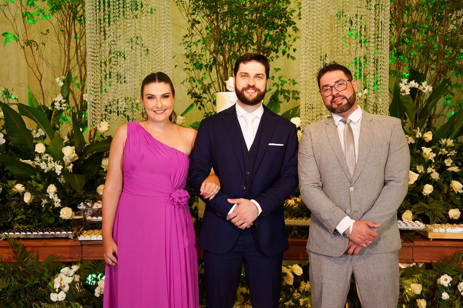 Three elegantly dressed guests pose together at a formal wedding reception with floral decor.
