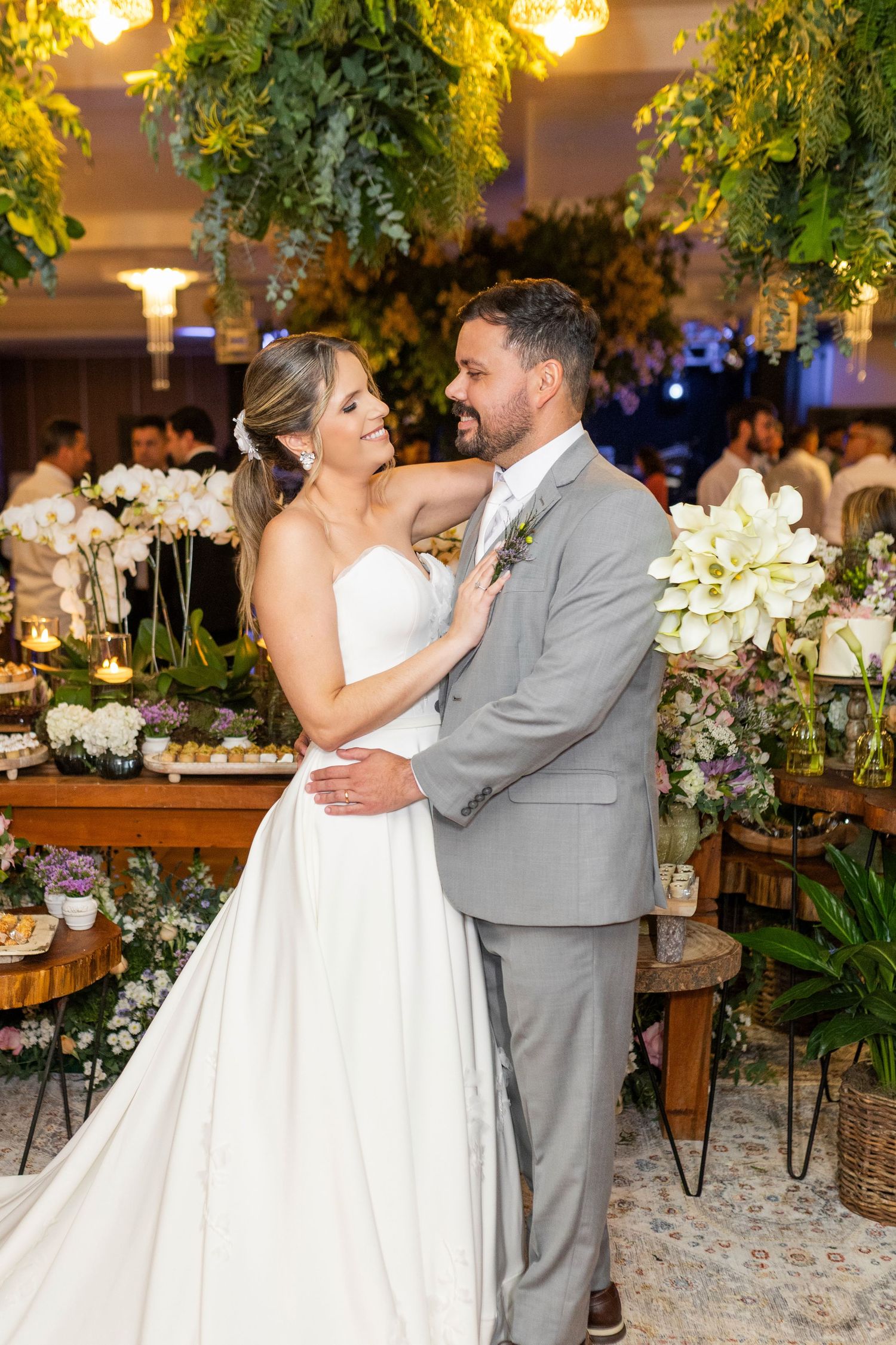 A bride in a white strapless gown and groom in a gray suit share a romantic moment at a floral wedding reception.