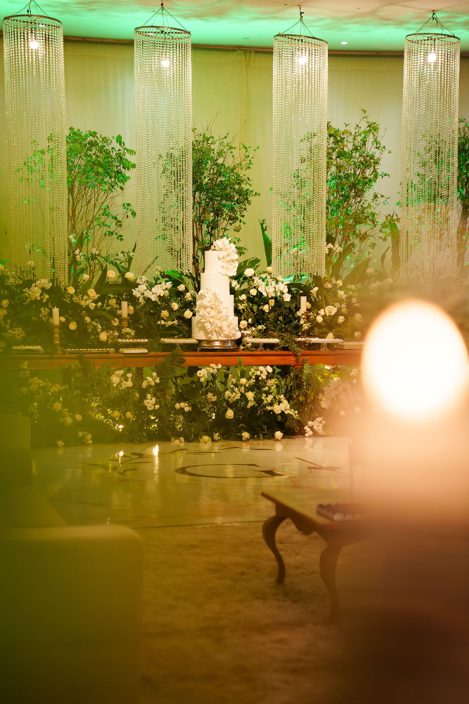 Elegant wedding reception with white tiered cake, lush greenery, white floral arrangements, and crystal chandeliers in warm lighting.