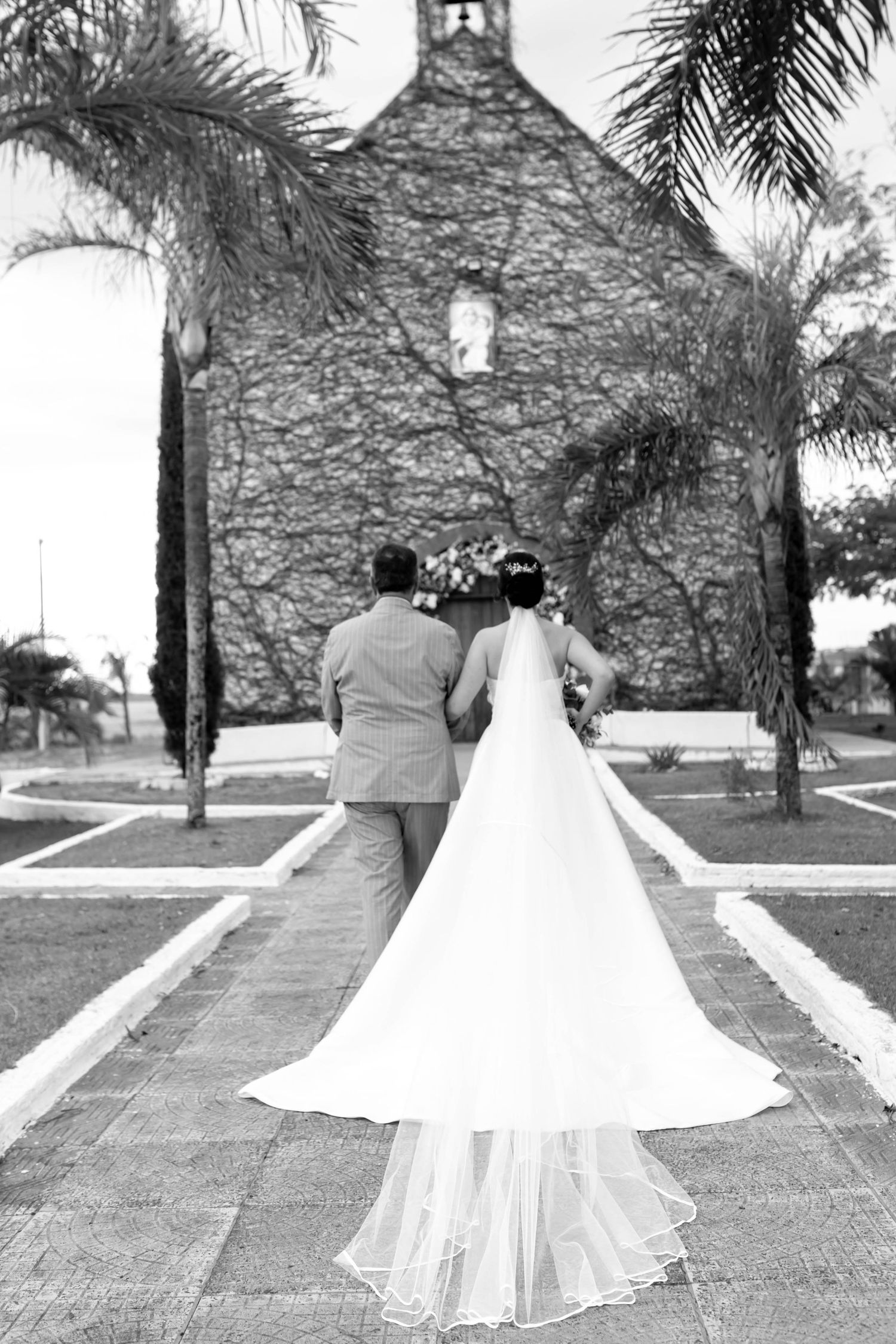 Black and white photo of bride and groom walking toward a vine-covered church, bride's long train trailing behind.