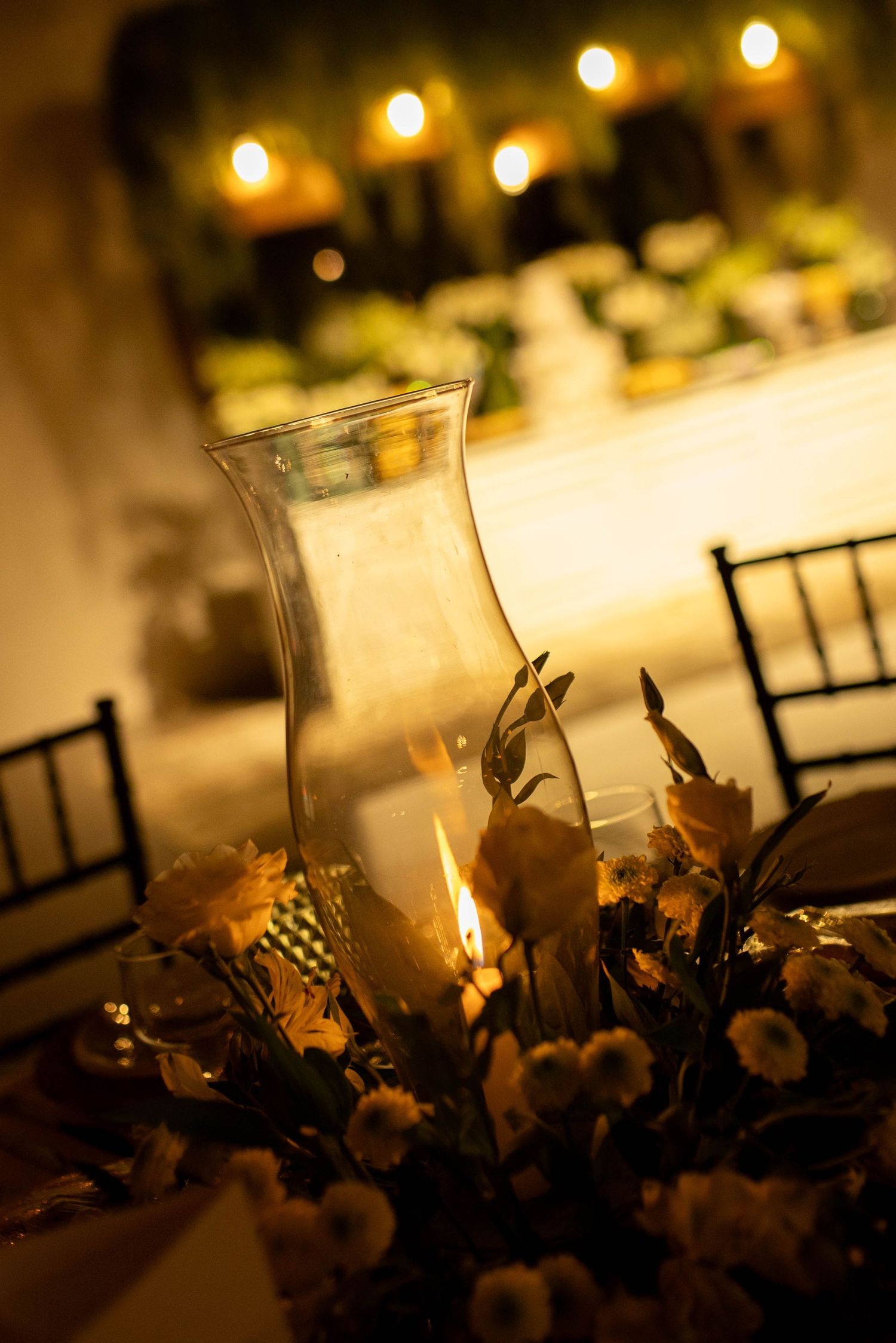 Elegant candlelit wedding reception table with glass hurricane vase, white flowers, and warm glowing lights in background.
