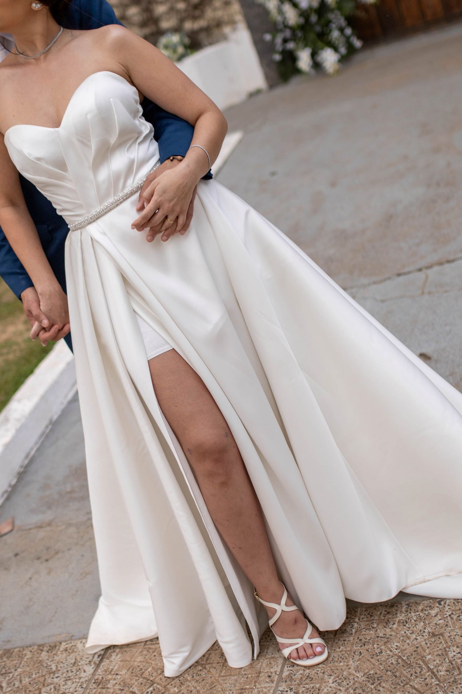 Bride wearing elegant strapless white satin wedding gown with thigh-high slit and white strappy heels outdoors.
