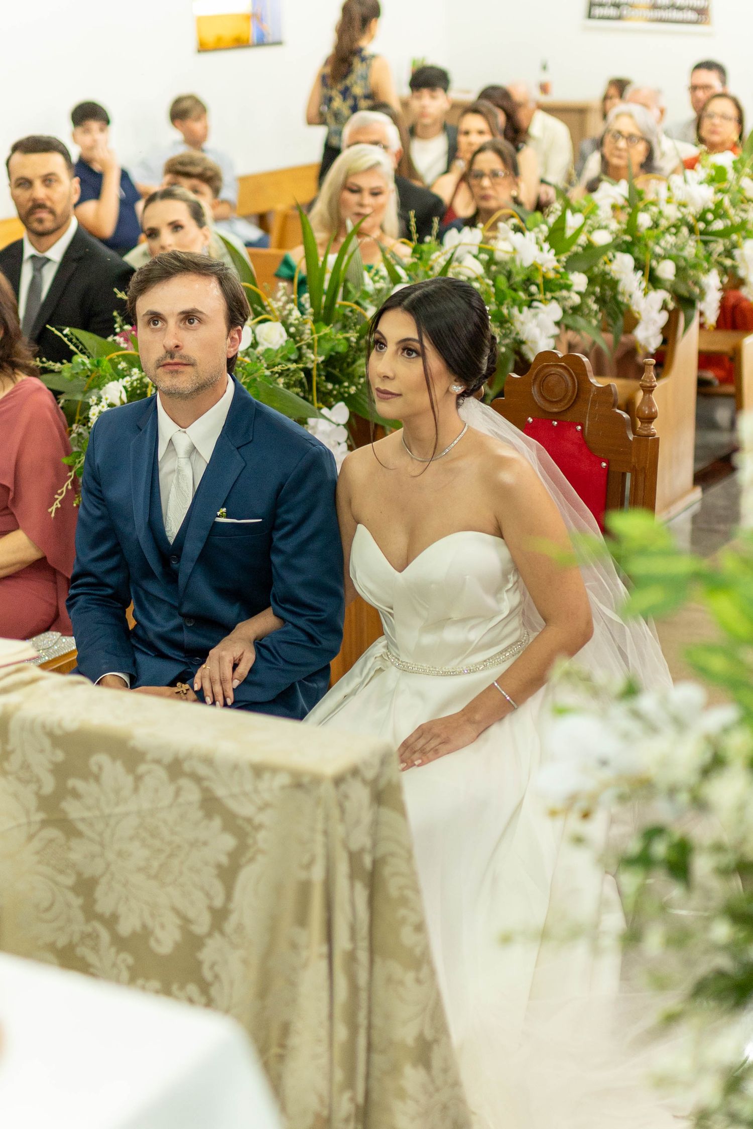Bride in white strapless gown and groom in navy suit seated at altar during church wedding ceremony with floral decorations.