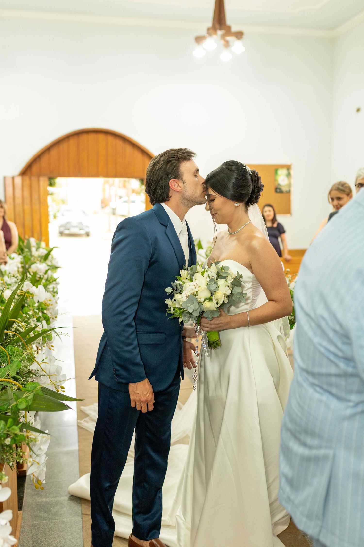 Groom in navy suit kisses bride holding white bouquet at wedding ceremony in elegant chapel.