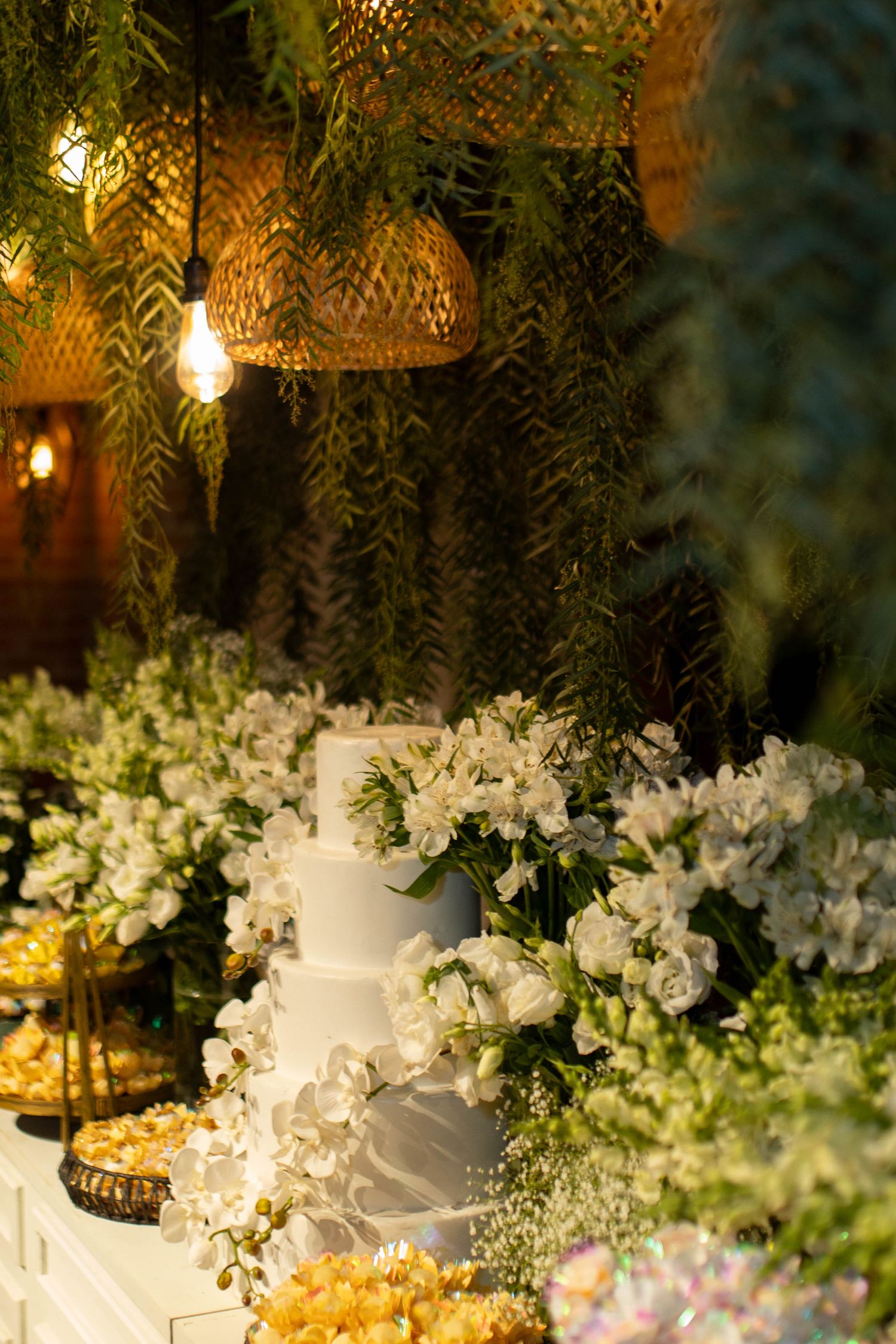 Elegant white tiered wedding cake surrounded by lush white floral arrangements under warm wicker pendant lights.
