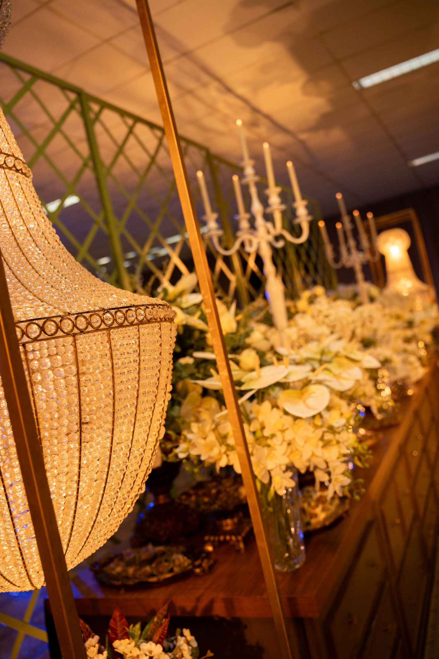 Elegant event decor with crystal chandelier, white floral arrangements, candelabra, and golden accents in a warmly lit venue.