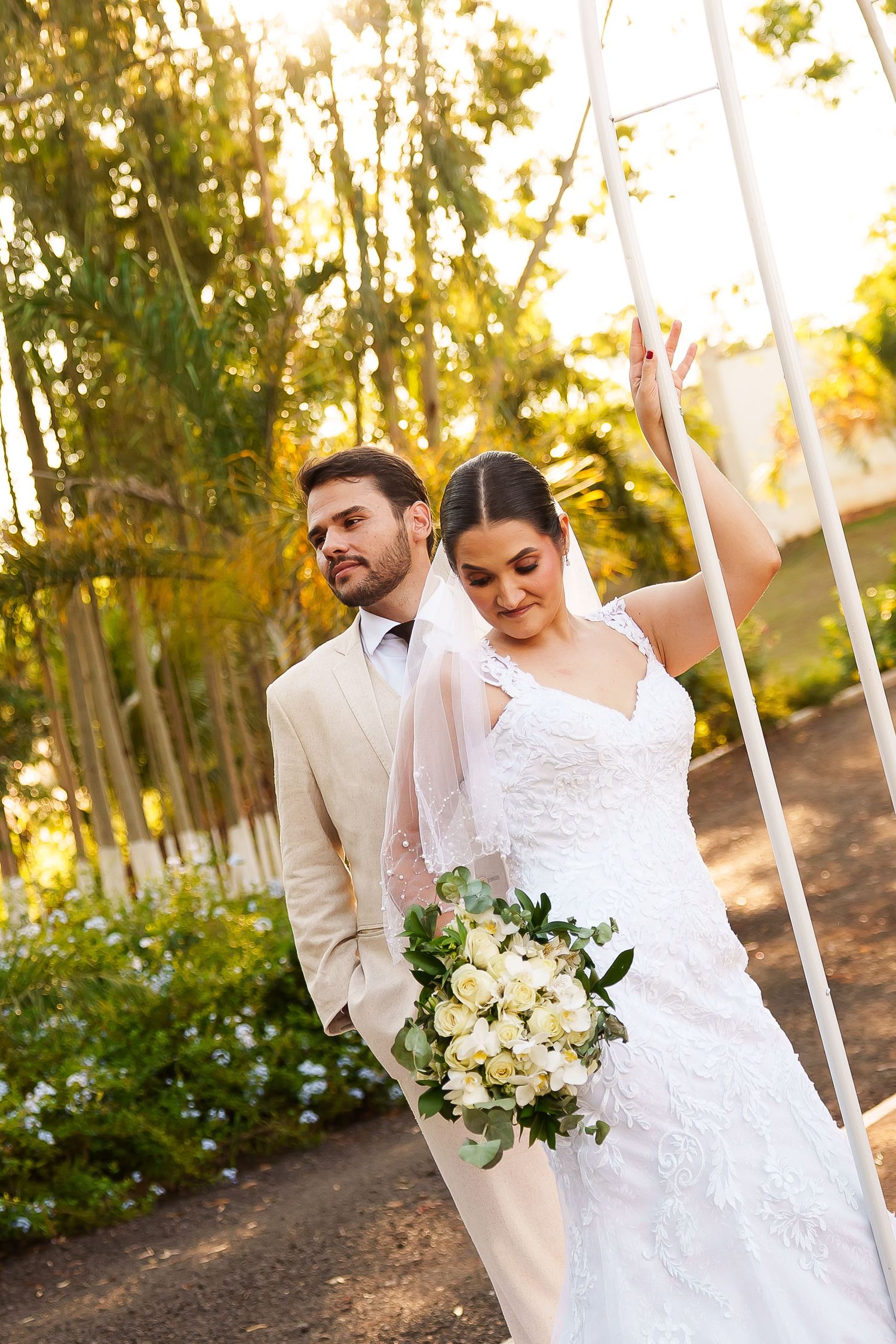 Bride in lace gown holding white bouquet stands with groom in beige suit near white arch in sunlit garden.