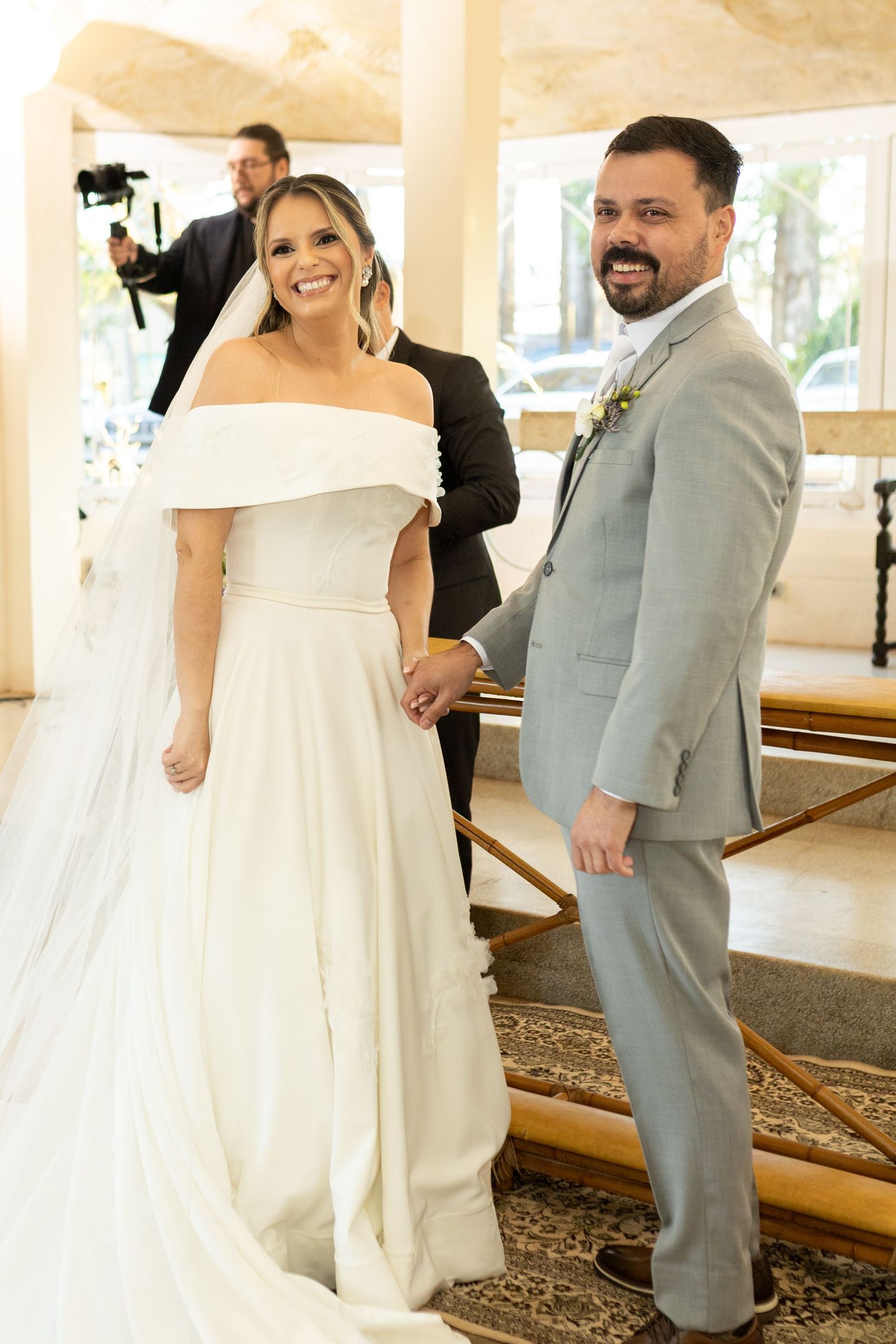 Smiling bride in off-shoulder white gown and groom in gray suit hold hands at their wedding ceremony.