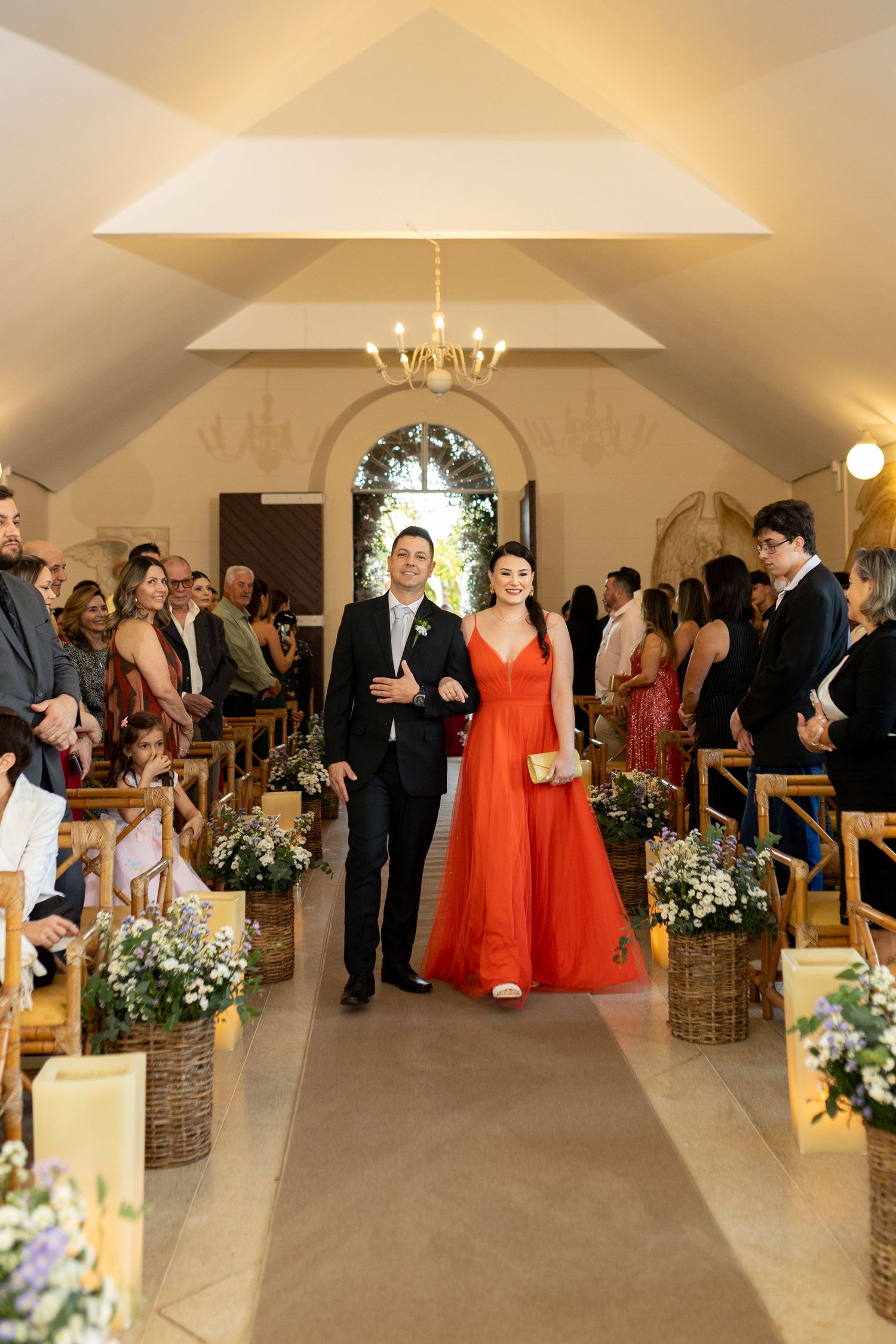 Smiling bride in orange gown walks chapel aisle with suited escort past seated wedding guests.