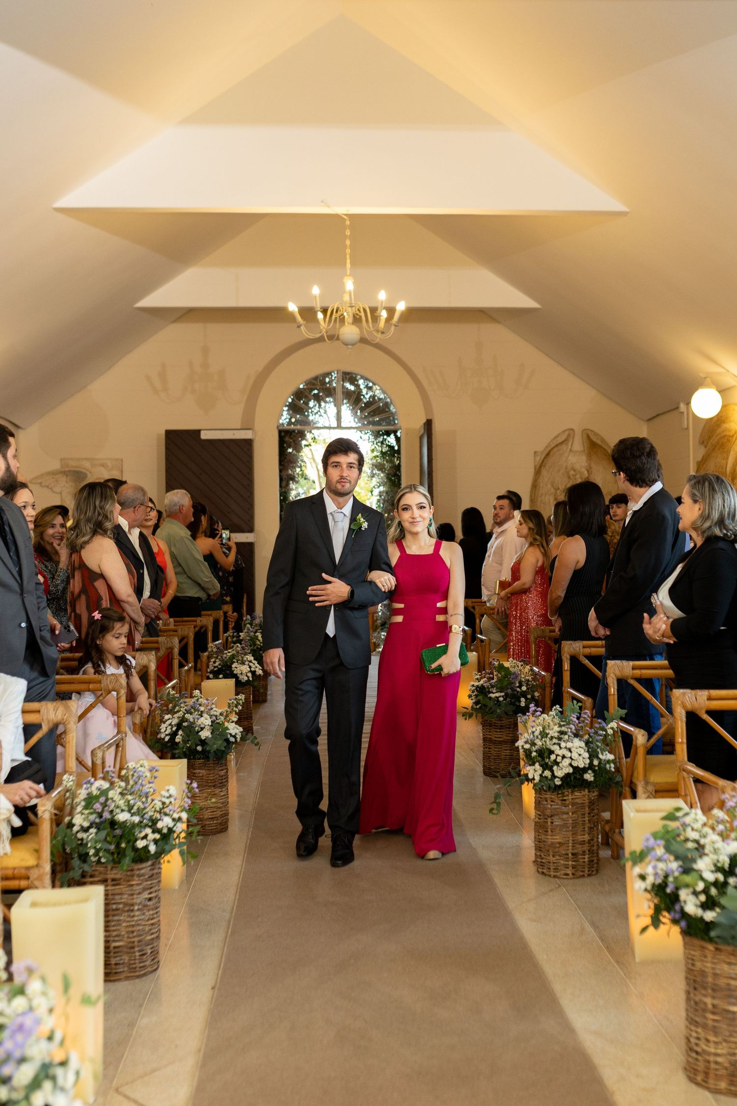 Man in suit and woman in red gown walk down floral-decorated church aisle at wedding ceremony.