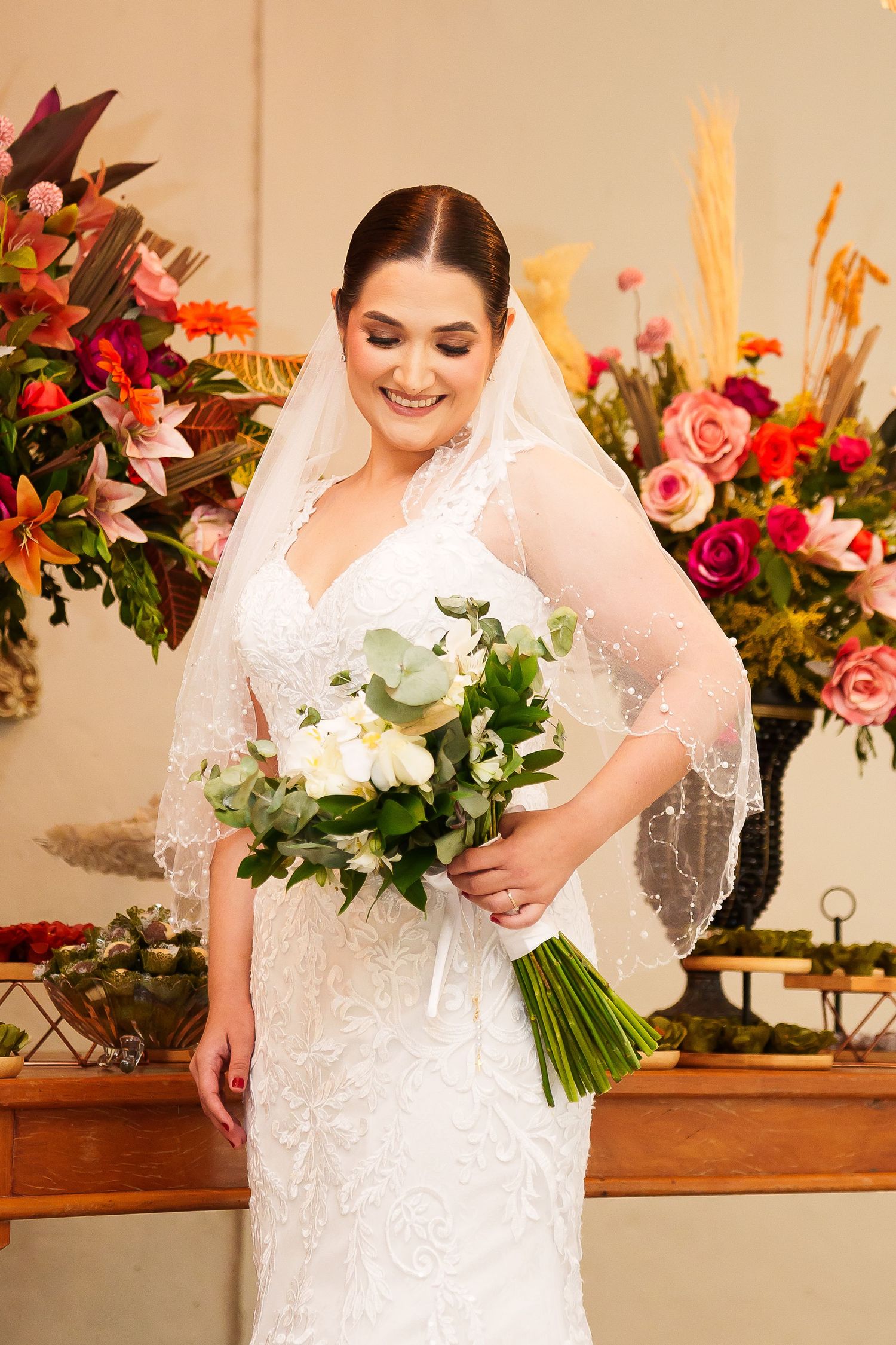 Smiling bride in white lace gown and veil holds bouquet of white flowers, standing before colorful floral arrangements.