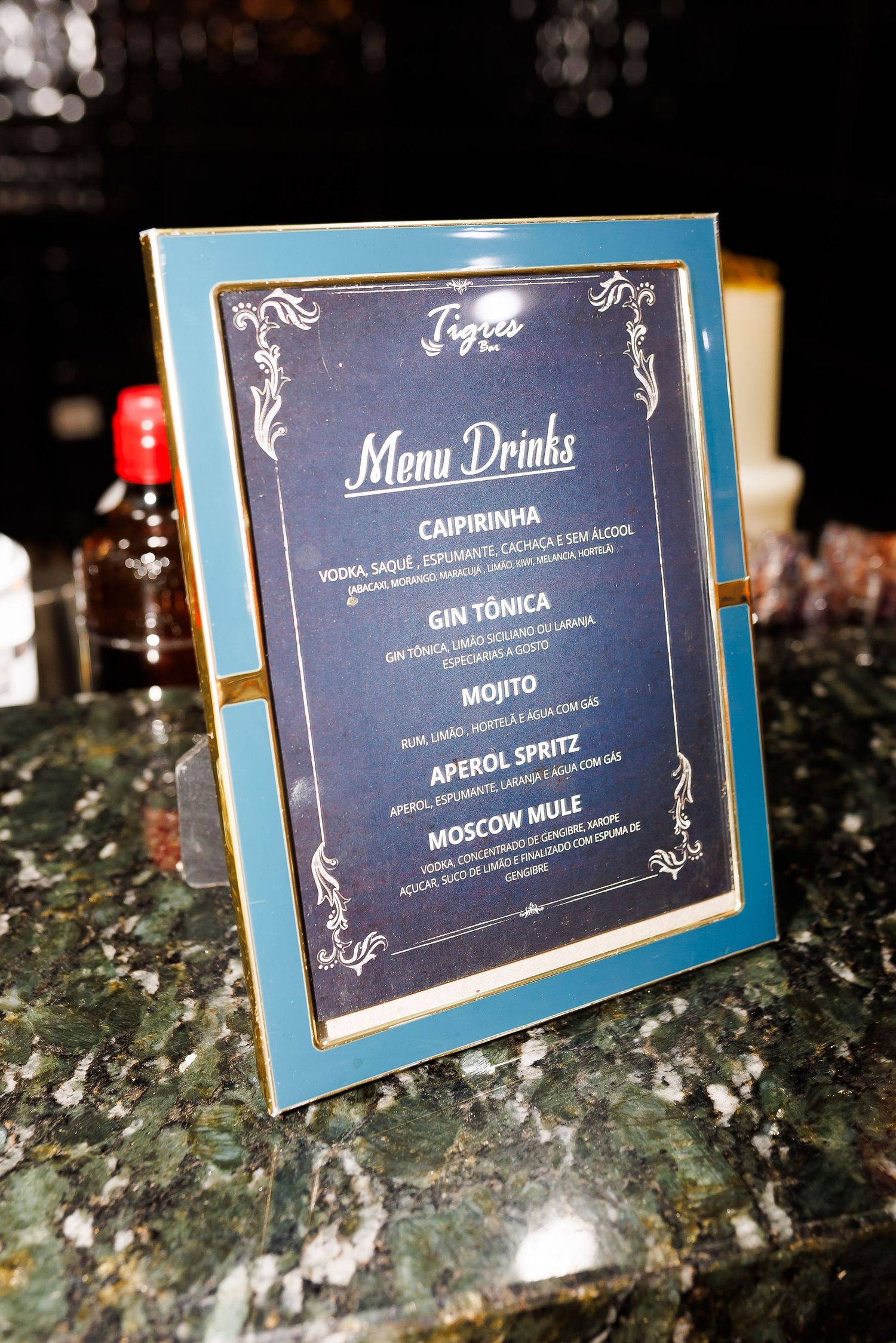 Elegant blue and gold framed cocktail menu displaying drinks like Caipirinha, Gin Tonica, Mojito, Aperol Spritz, and Moscow Mule.
