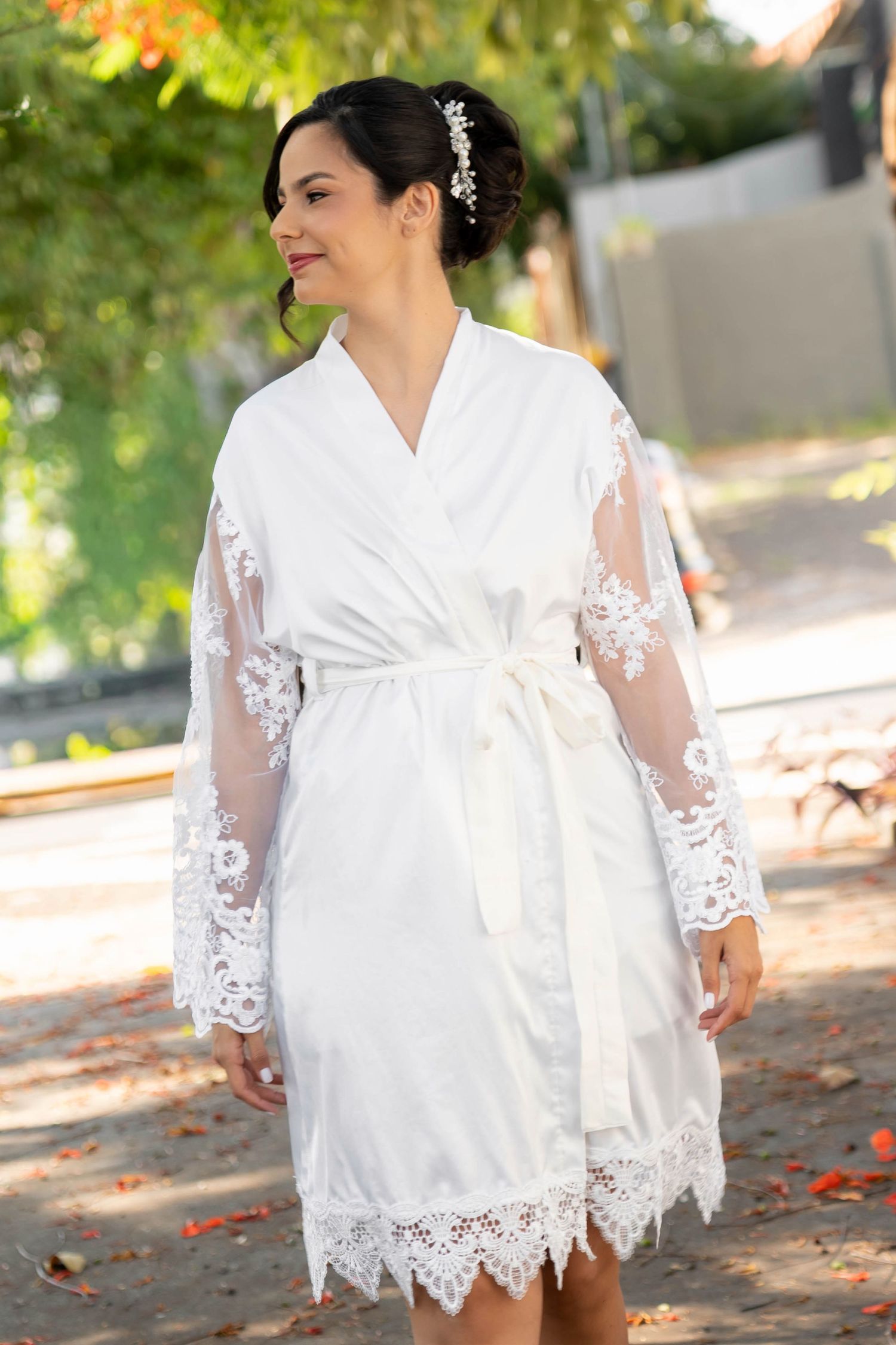 Bride wearing a white lace-trimmed robe with sheer sleeves and floral hair accessory, smiling outdoors.