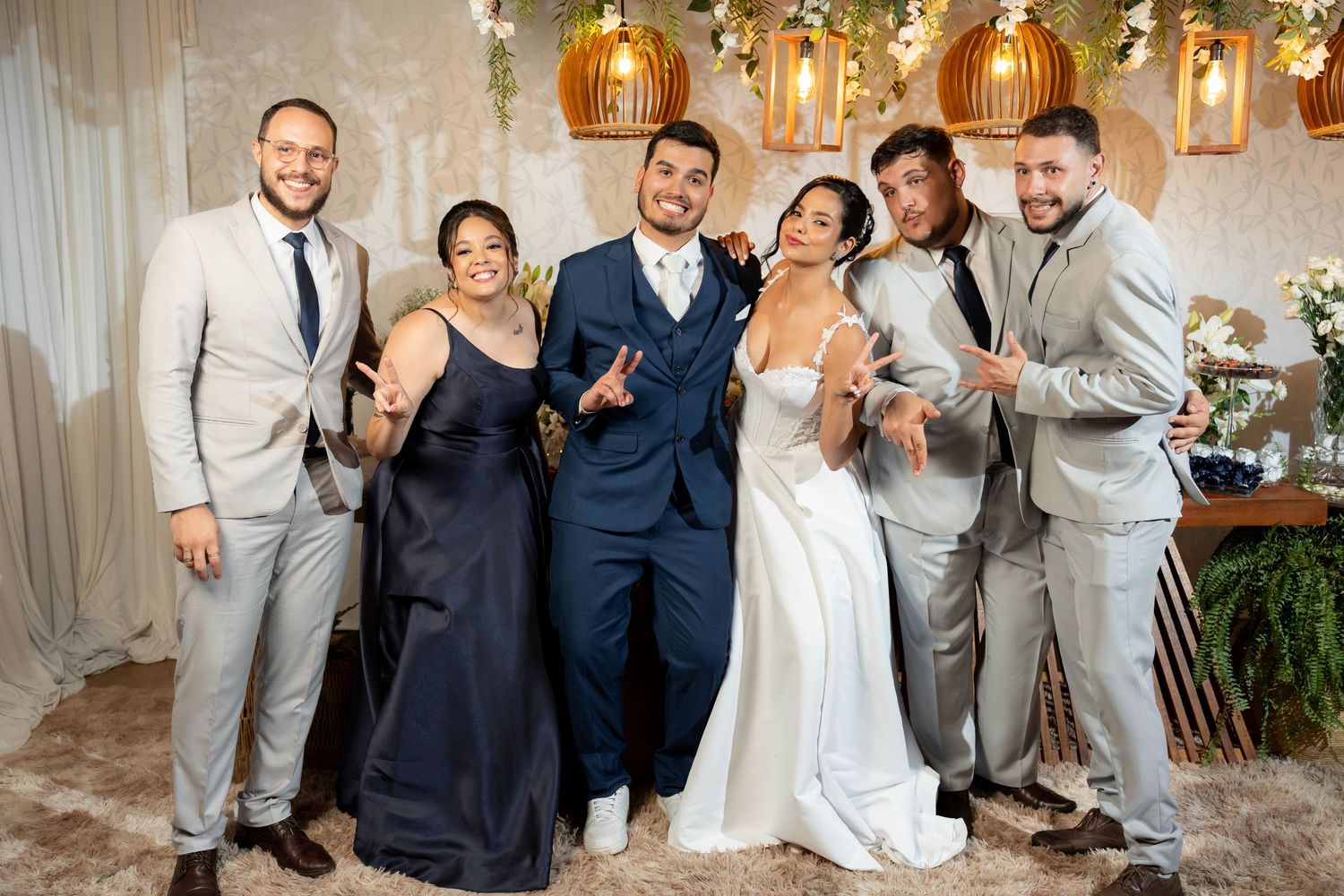 A joyful wedding group photo featuring a bride in white, groom in navy, and four guests in gray suits and a black gown.