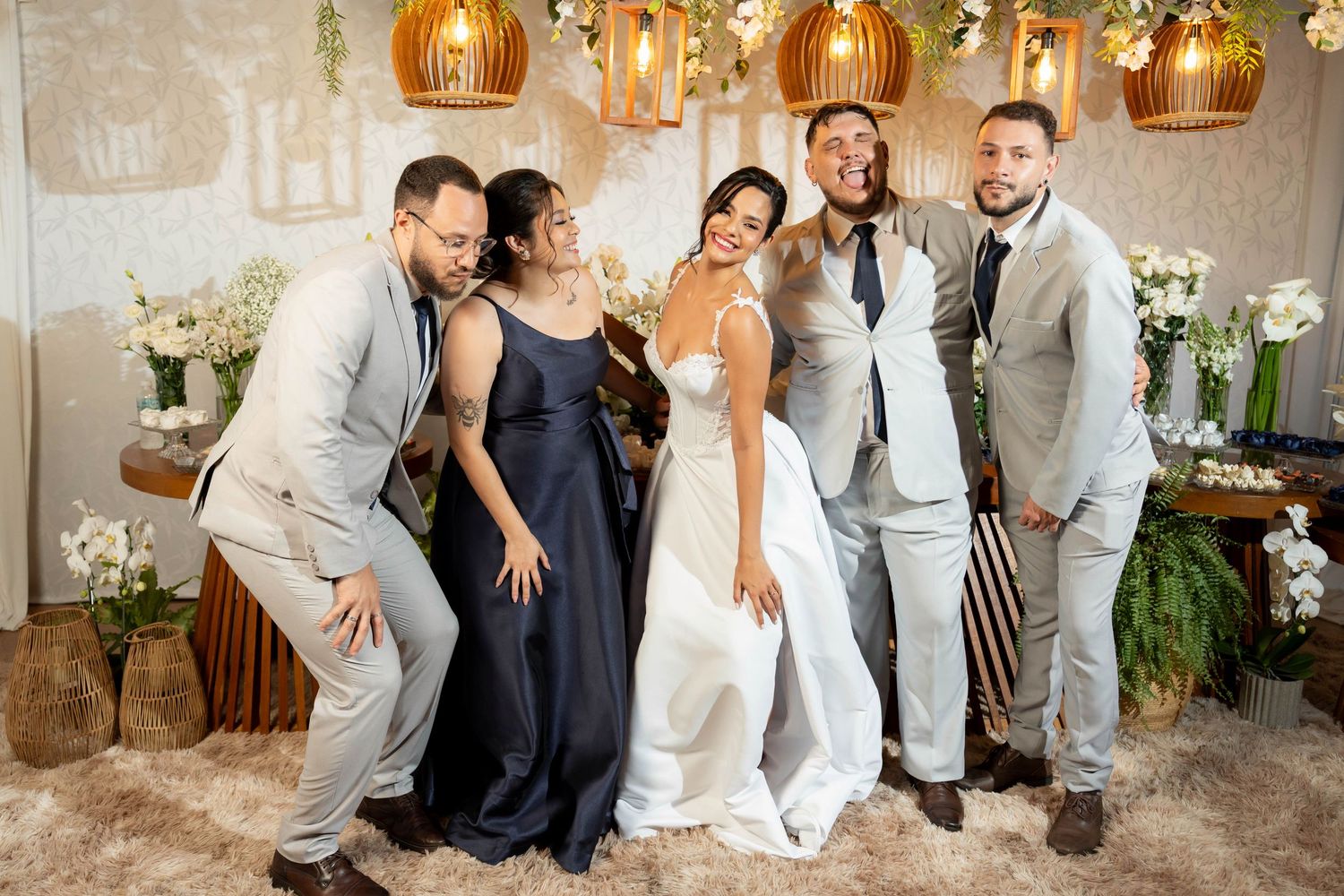 A joyful wedding group photo featuring a bride in white and guests in formal attire, posing playfully amid floral decor.