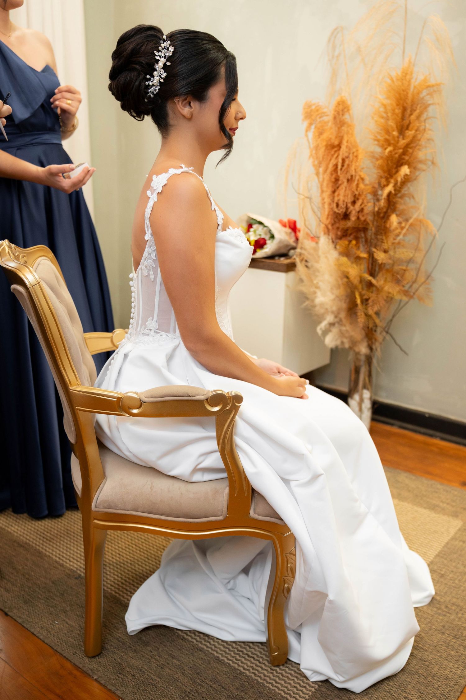 Bride in white lace gown seated in gold chair, hair up with floral accessory, bridesmaid in navy dress beside her.