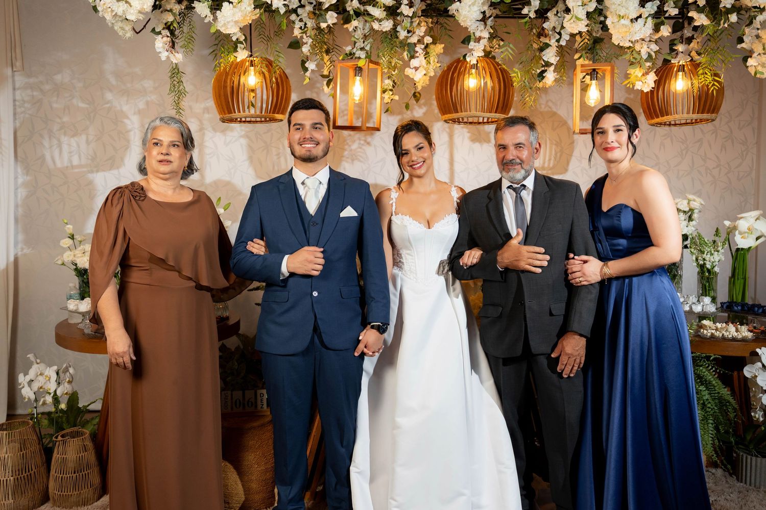 Wedding party of five posing indoors, bride in white gown, groom in navy suit, guests in blue and brown dresses.