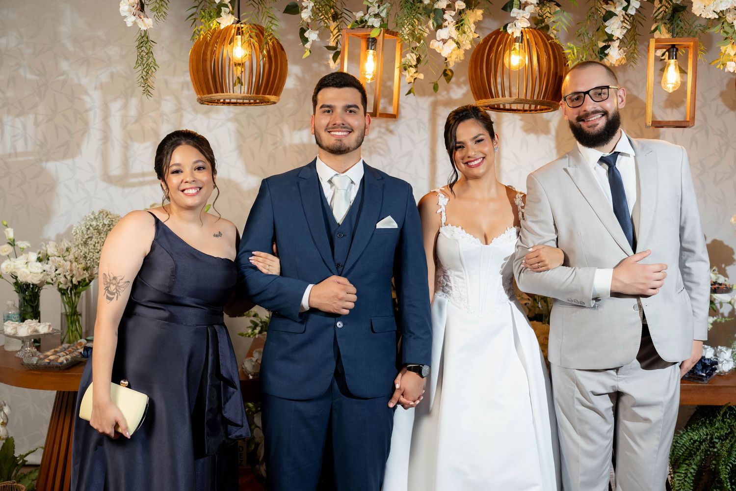 Wedding party of four posing indoors, bride in white gown, groom in navy suit, guests in black dress and gray suit.